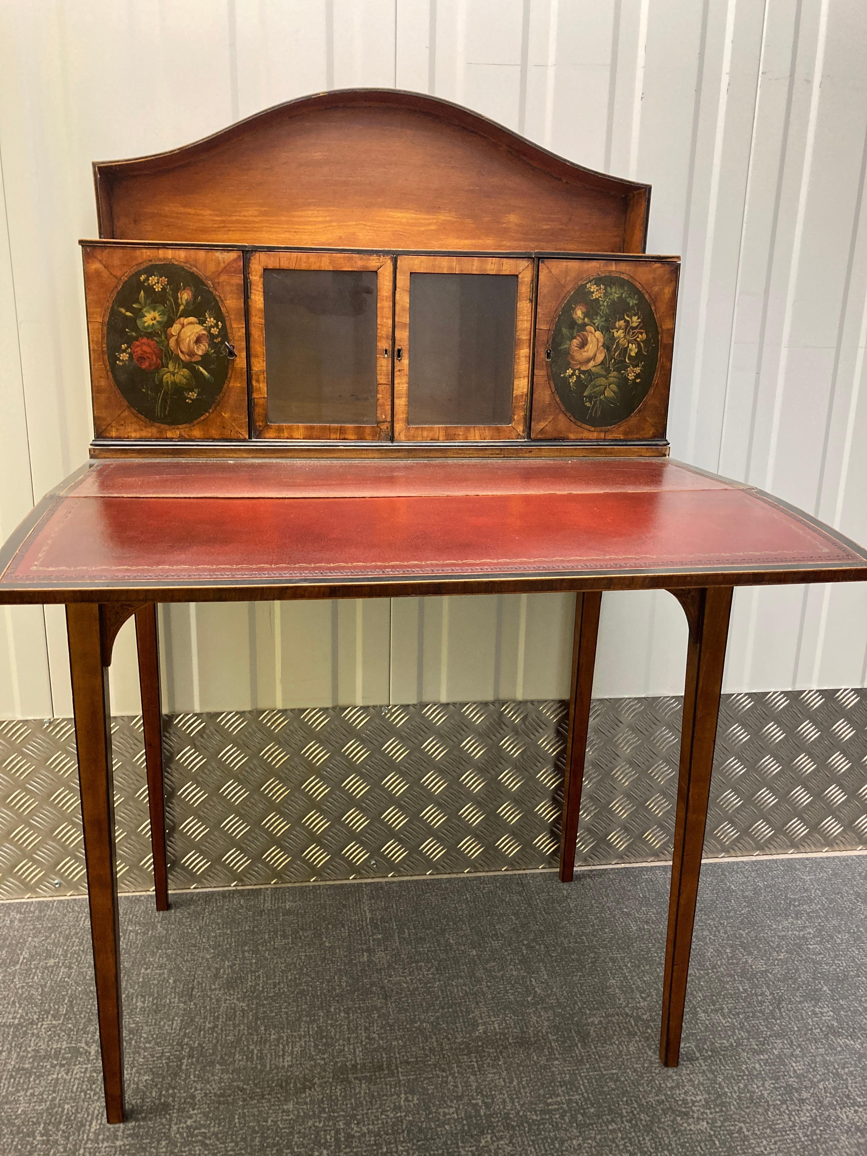 A Satinwood Bonheur du Jour with painted panels and glazed doors, maroon leather writing fold, ebony inlaid square tapering legs and painted brackets. After a design by Hepplewhite. English circa 1790  