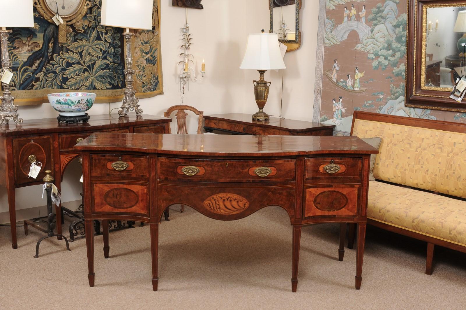 An English George III serpentine mahogany and satinwood sideboard featuring 2 cellarette drawers, center silver drawer and shaped apron below with shell inlay ending on tapered legs with spade feet.
