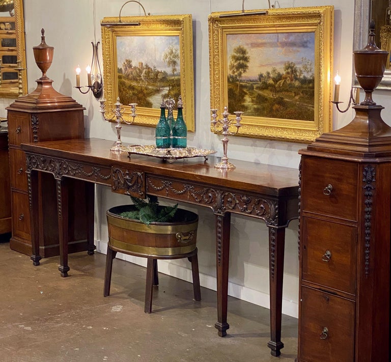 A rare and important period English serving table or console server with matching lead-lined dining room pedestals of mahogany from the George III Era.
Featuring handsomely carved features of acanthus foliage and husks, interspersed with urns and