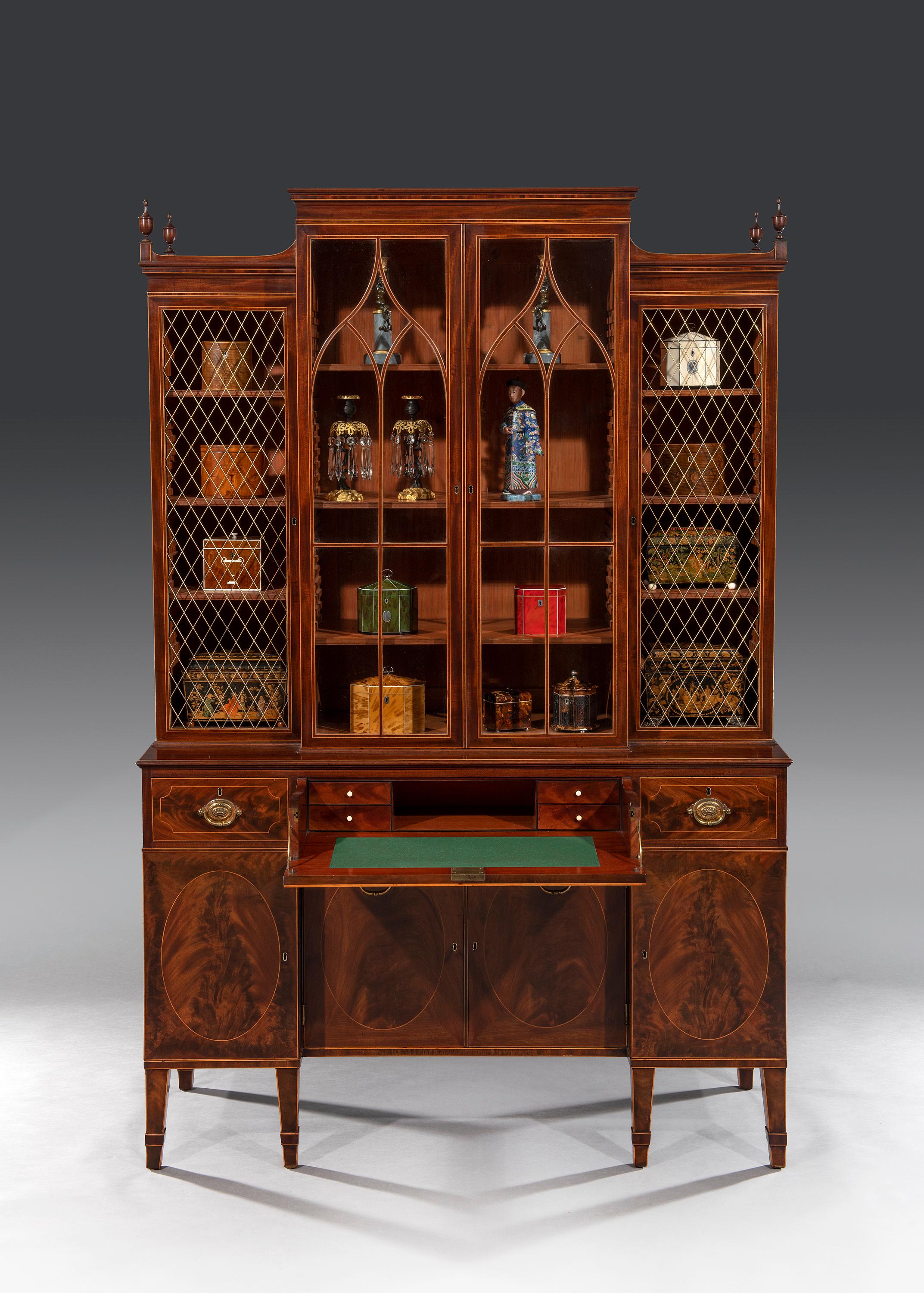 The cabinet has a shaped top to the centre and carved urns to the top of each side above rectangular glazing bars. The lower section has a fitted central writing drawer with a recessed cupboard below. The sides are fitted with single drawers above