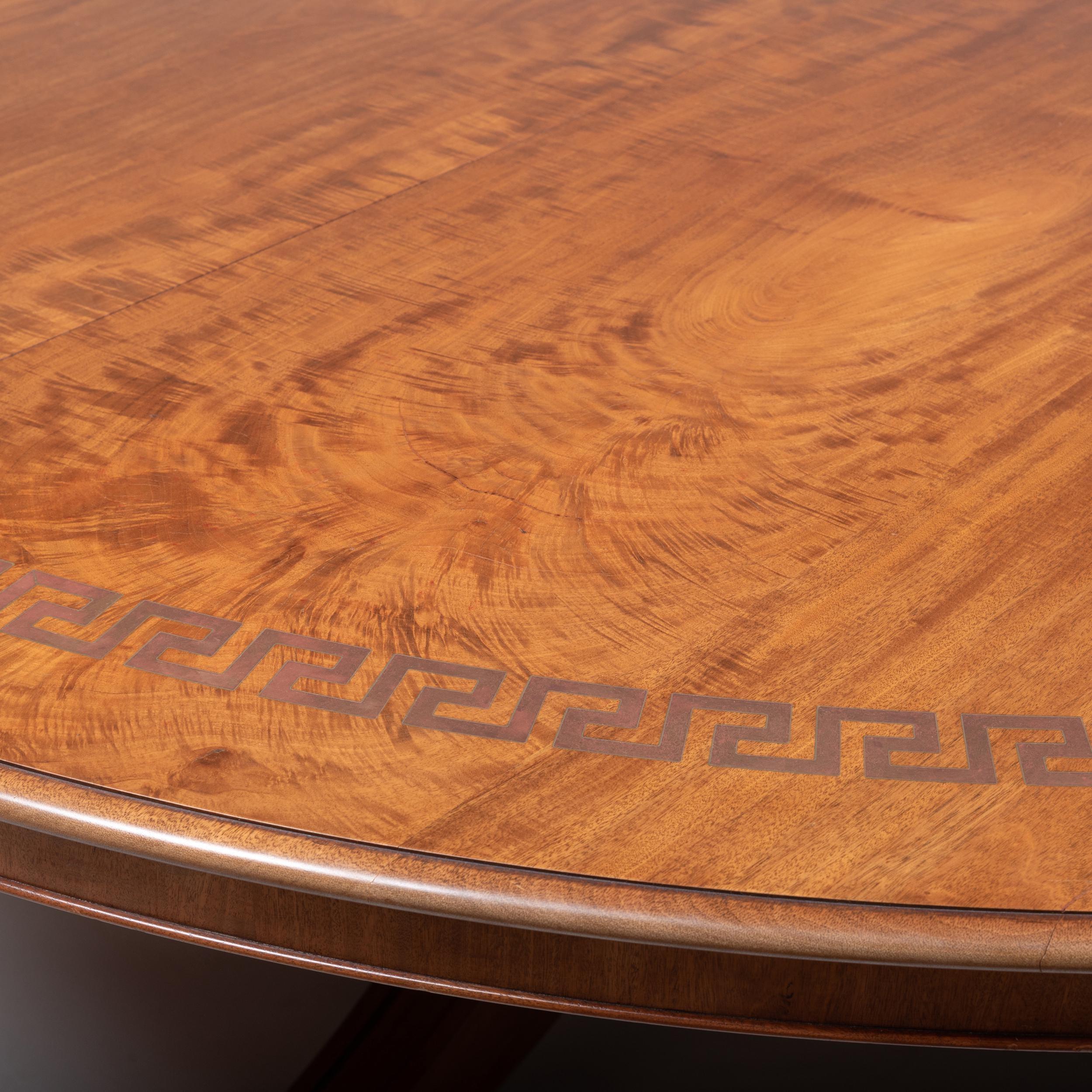 Round mahogany dining table in George III style with Greek key design at perimeter in brass inlay. Table sits on pedestal base with four curved legs ending in brass caps.