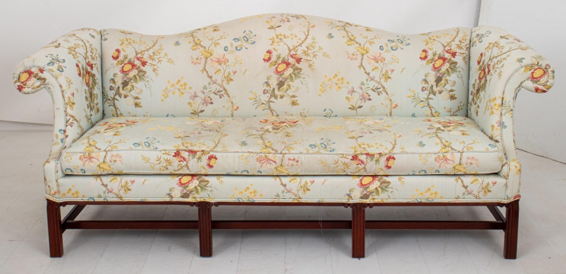 George III style camelback sofa upholstered allover in multicolored silk lampas, with arched back and scrolling arms above a drop in seat, on bracket legs conjoined by stretchers. 

Dimensions: 34