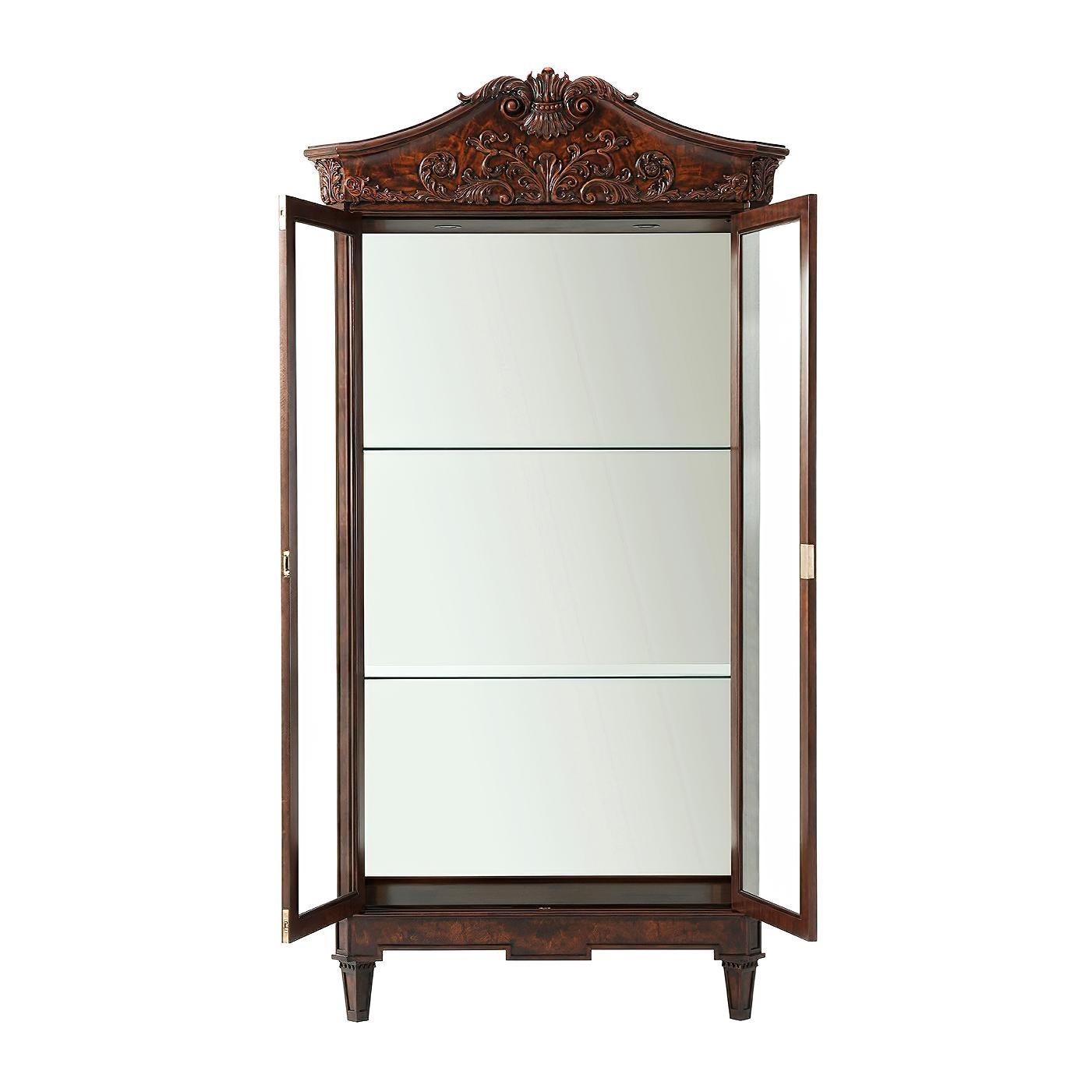 A fine George III style flame mahogany, Morado banded and Movingue display cabinet, the arched hand carved mahogany and flame mahogany veneered cornice with a shell crest, above two glazed doors and sides, enclosing a lit interior with a mirror back