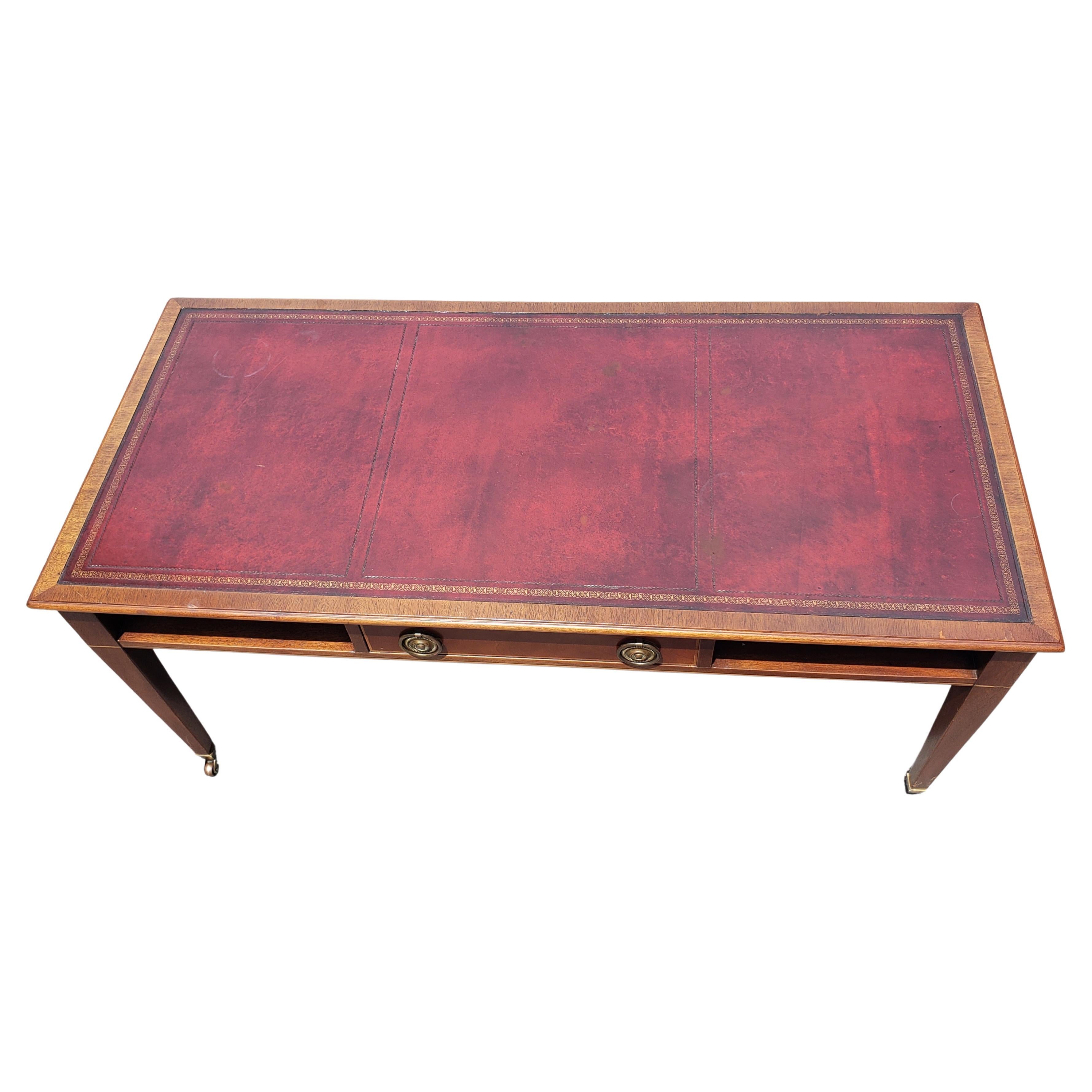 A beautiful  Mahogany Maroon Leather Inset Top Coffee Table on Wheels in the George III Style. Table features Banded top edges, original wheels and one top center drawer with two side storage shelves.  Table is in good vintage condition and measures