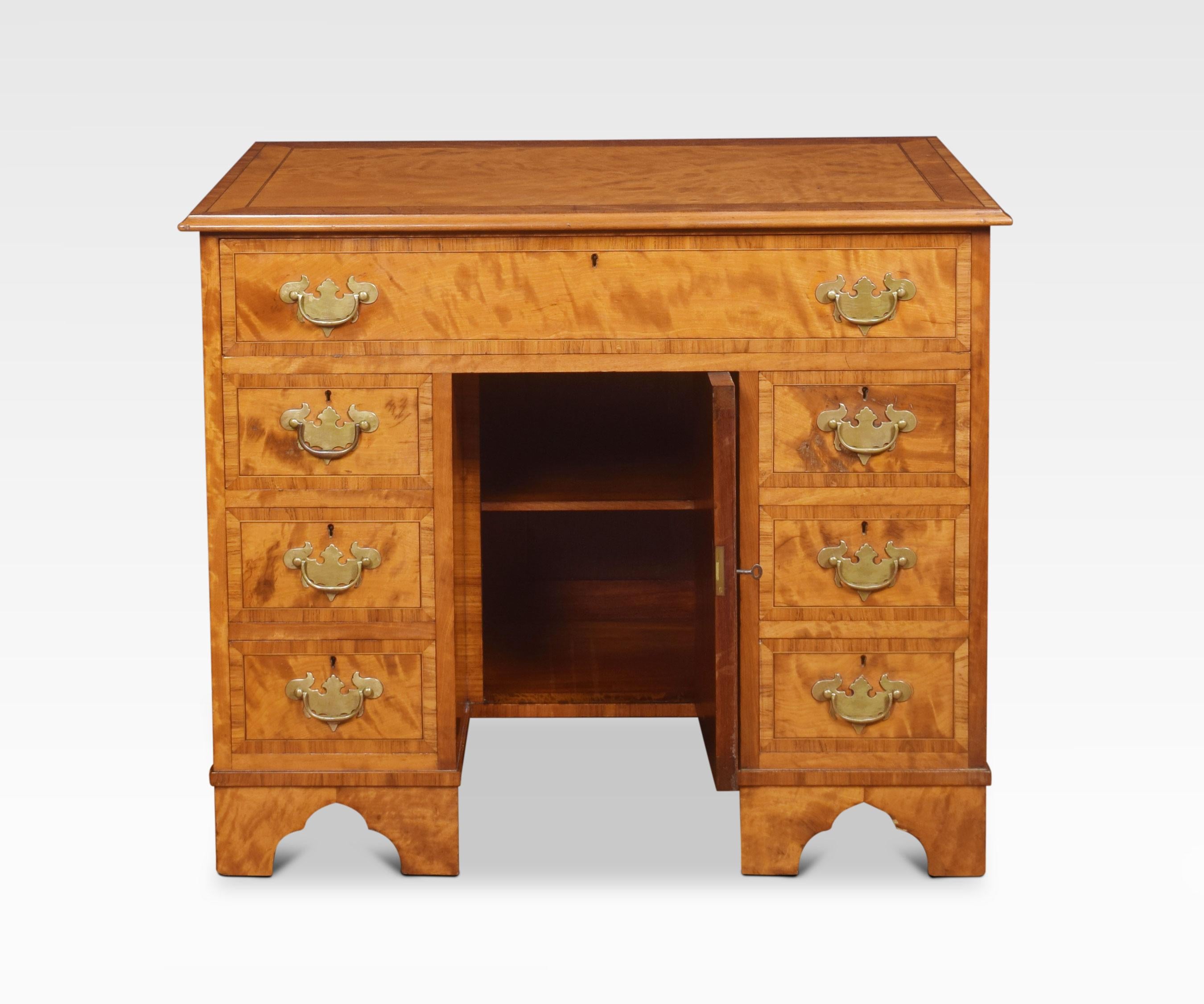 George III style satinwood kneehole desk. The rectangular top with a crossbanded edge above a large freeze drawer. To the recessed cupboard door with similar crossbanded detail. flanked to each side by three drawers with brass handles. All raised up