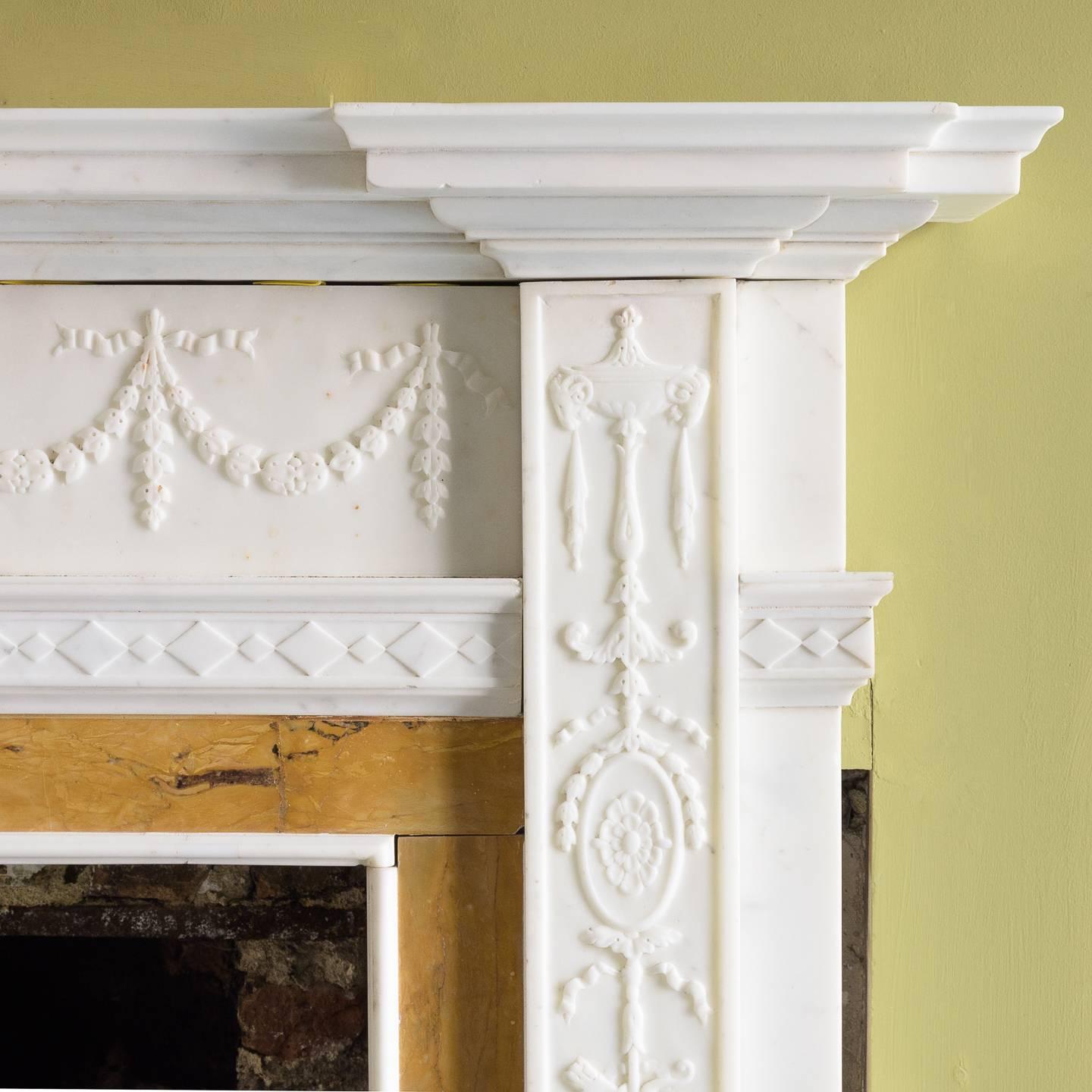 A George III style statuary and sienna marble chimneypiece, circa 1900 in the neoclassical style, the inverse breakfront moulded shelf above frieze with floral swags centred by tablet depicting playing putti and a ram, the pilaster jambs with