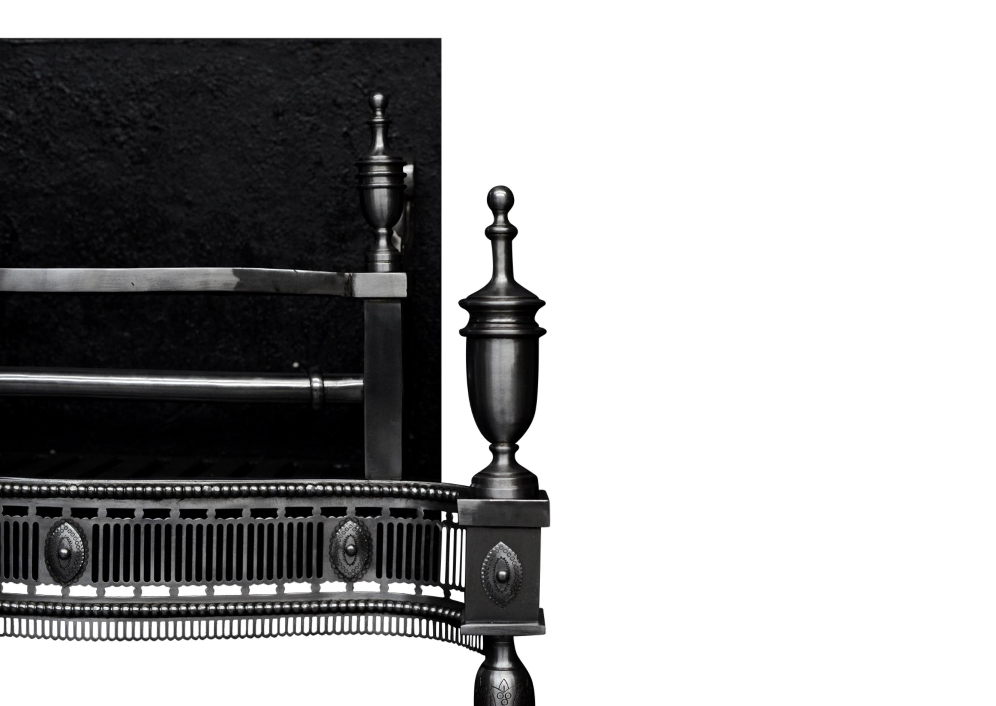 A Georgian style steel fire grate. The serpentine shaped fret with flutes, beading and engraved paterae. The four finials of urn shape, sat on square plinths with matching paterae, and shaped legs below. Scrolled side returns leading to square plain