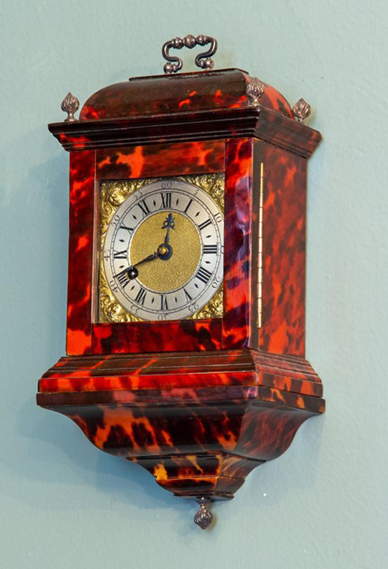 An ultra rare Georgian miniature tortoiseshell bracket timepiece travel clock, caddy top with white metal swing handle and flame finials, the 7cm square brass dial with silvered chapter ring with Roman numerals and mask spandrels, single fusee chain