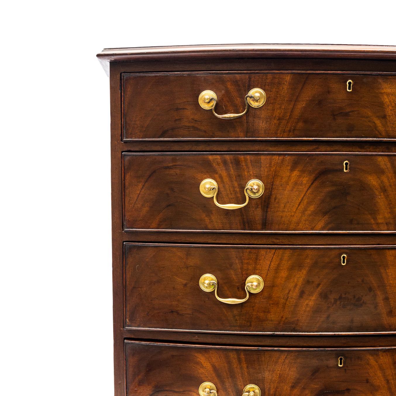 A George IV Period Bowfront Chest in Figured mahogany, with four graduated and cock-beaded drawers, the top with a molded edge, on a shaped apron base and graceful splayed feet (original), with hand-forged brass bail pulls (original), with oak