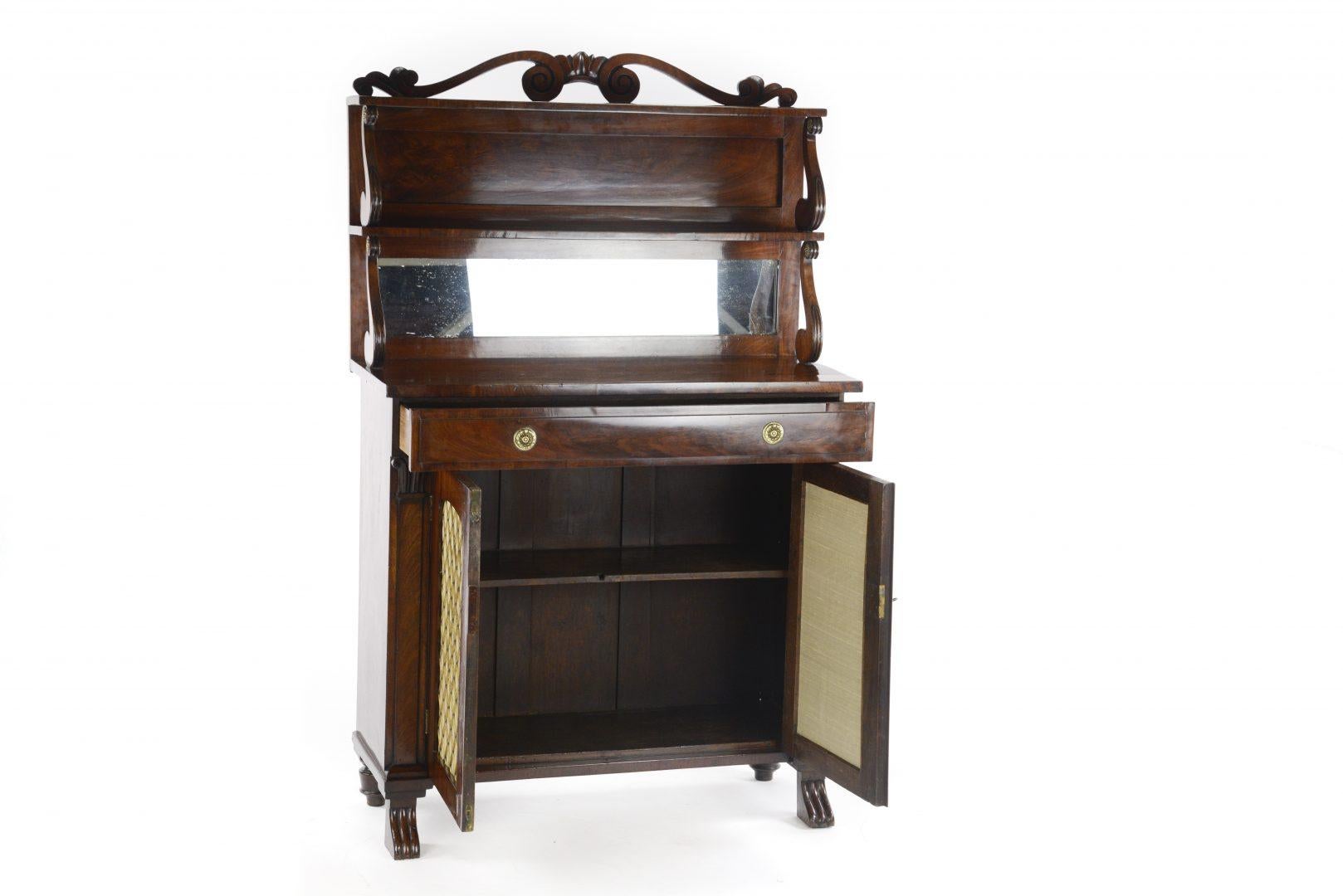 A modest George IV mahogany chiffonier dating to circa 1825, having an upper section with two mahogany shelves with a gallery supported on finely carved brackets, two rectangular mahogany doors with brass grilles and silk lined panels and having