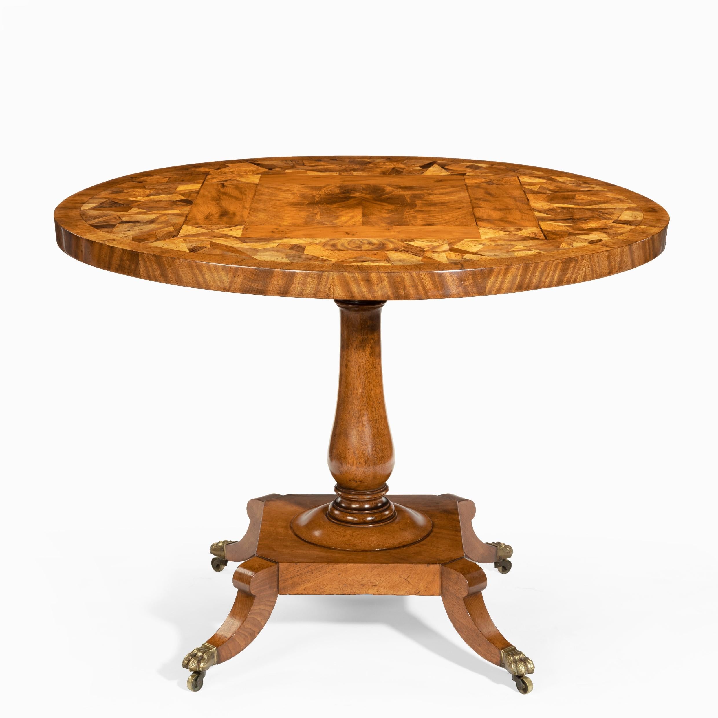 A George IV tilt-top specimen table, the circular top inlaid with a central square of flame mahogany within a yew-wood frame all surrounded by a patchwork of exotic timbers, the turned mahogany baluster support raised on a square base and four