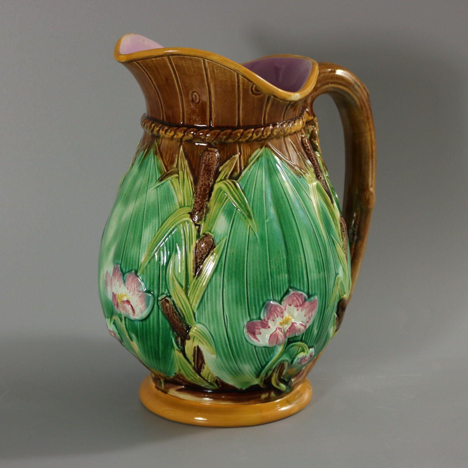 George Jones Majolica jug/pitcher which features green leaves, bulrushes/cattails and water lilies. Brown ground version. Colouration: brown, green, pink, are predominant. Bears a pattern number, '1800 T'.