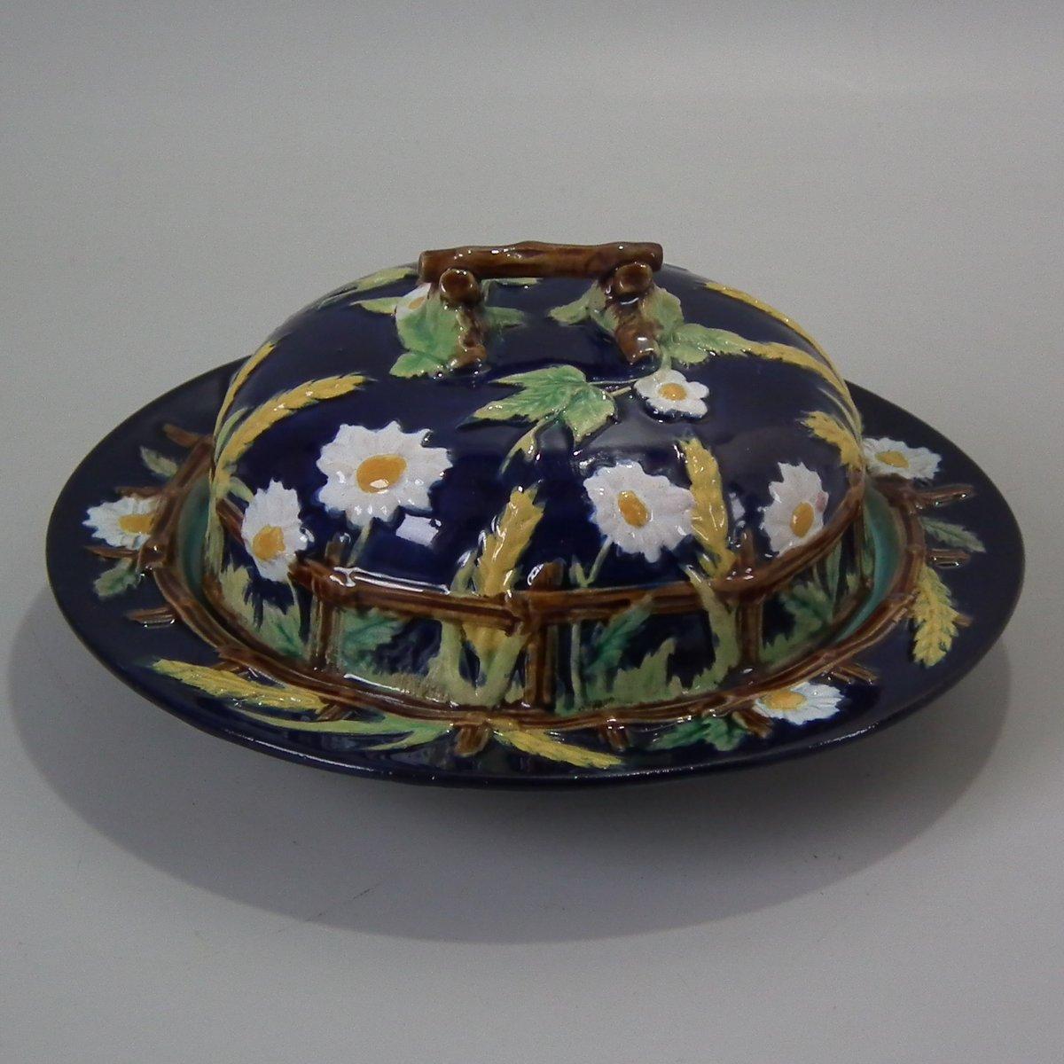George Jones Majolica muffin dish with cover which features a picket fence, daisies, corn, brambles and a twig handle. Cobalt blue ground version. Coloration: white, ochre, green, are predominant. Impressed and painted marks to the reverse.