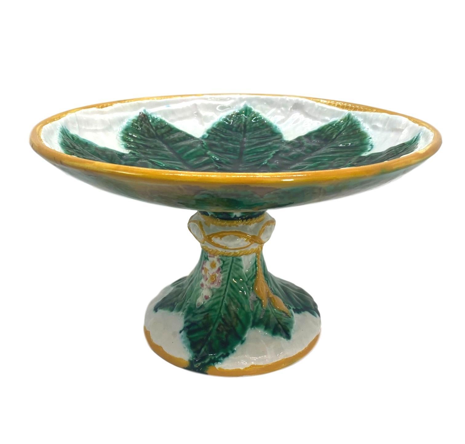 George Jones Majolica elevated comport, with a central green glazed horse chestnut leaf on a relief- molded white napkin, the border glazed in ochre, the pedestal wrapped with an ochre tassel, with horse chestnut leaves and blossoms, English, circa