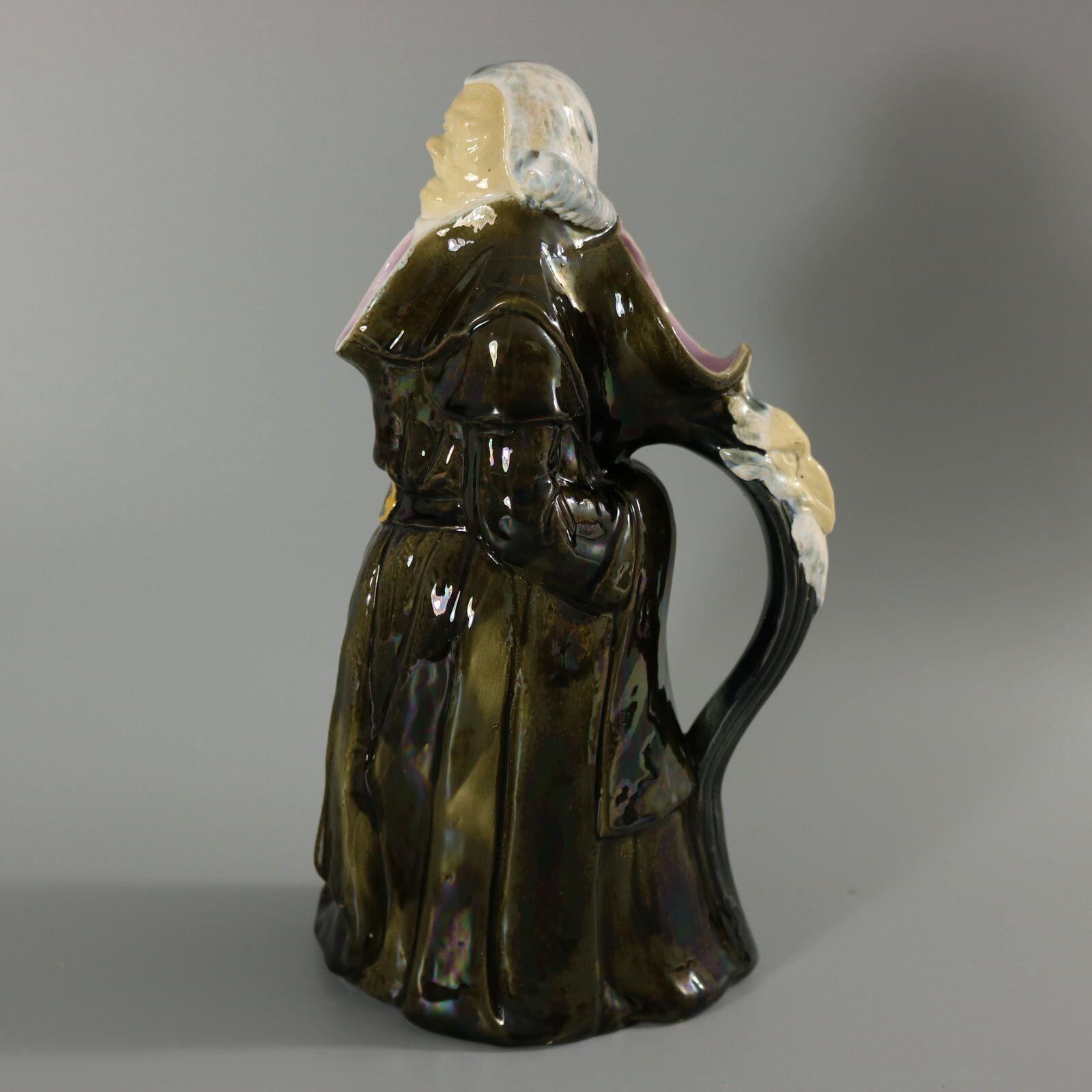George Jones Majolica jug/pitcher which features a monk, standing with hands behind his back. His belt has year 1750 inscribed. Goat head on handle. Colouration: grey, white, pink, are predominant. Bears a pattern number, '272?'.