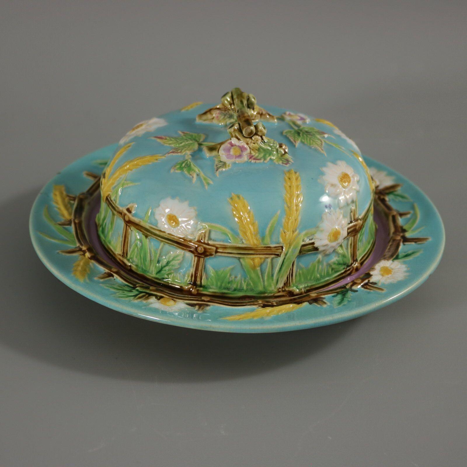 George Jones Majolica muffin dish and cover which features a picket fence, daisies, corn, brambles and a twig handle. Turquoise ground version. Colouration: turquoise, green, yellow, are predominant. Bears a pattern number, '1111'.