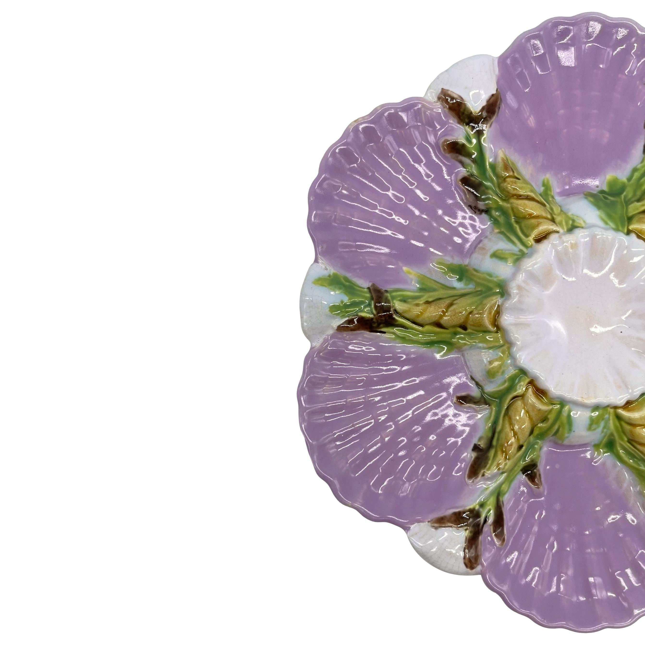 George Jones Majolica Oyster Plate, of hexifoil design, the six wells molded as scalloped seashells glazed in pastel lilac/pink, the wells separated by shells and seaweed, surmounted by a central scalloped shell forming the sauce well and glazed in