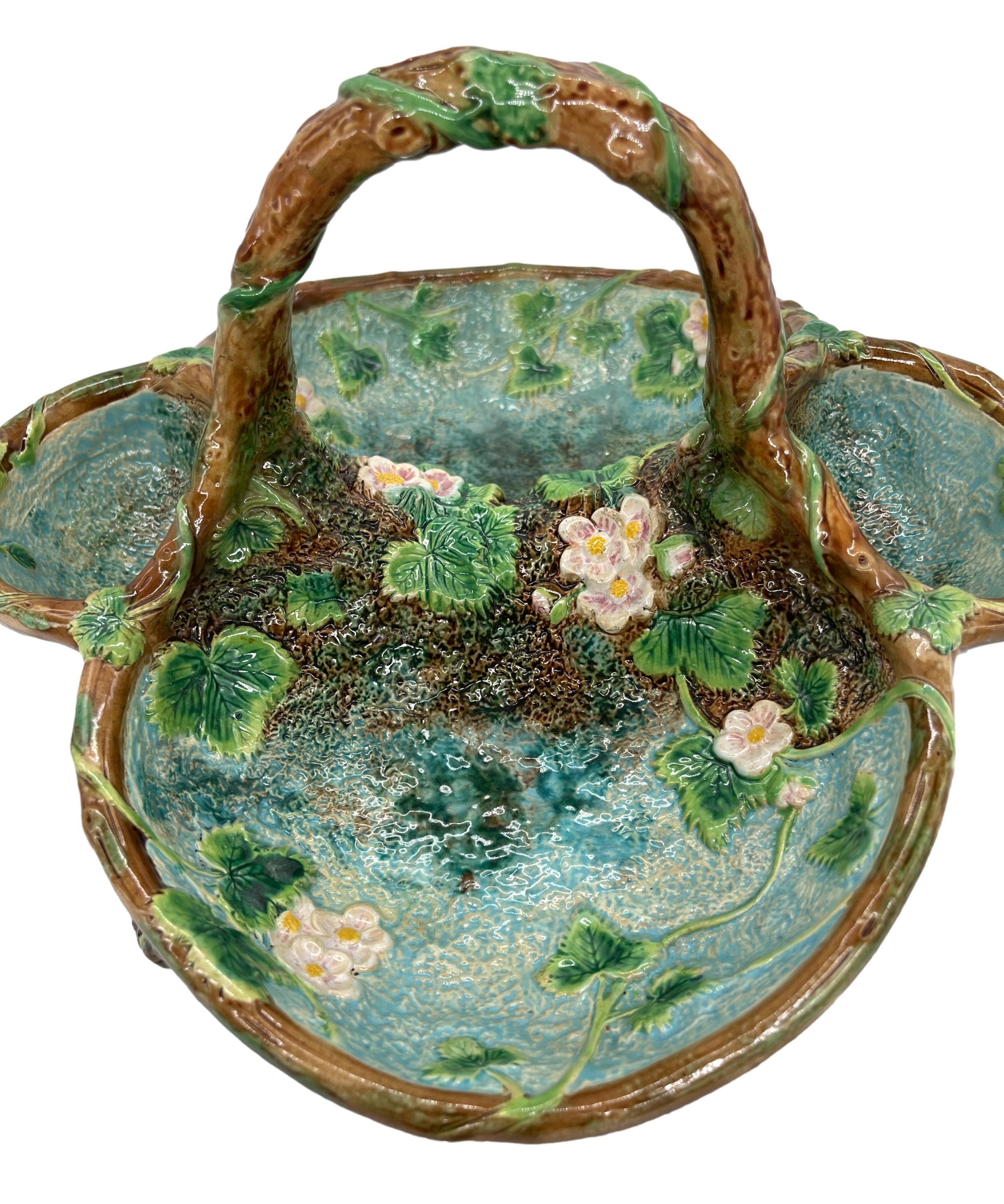 George Jones Majolica Rustic Strawberry Server, the quatrefoil basket naturalistically molded with blossoming strawberry plants and tendrils on a turquoise and mossy bark ground, the branch-form handle terminating in molded mossy pools to either