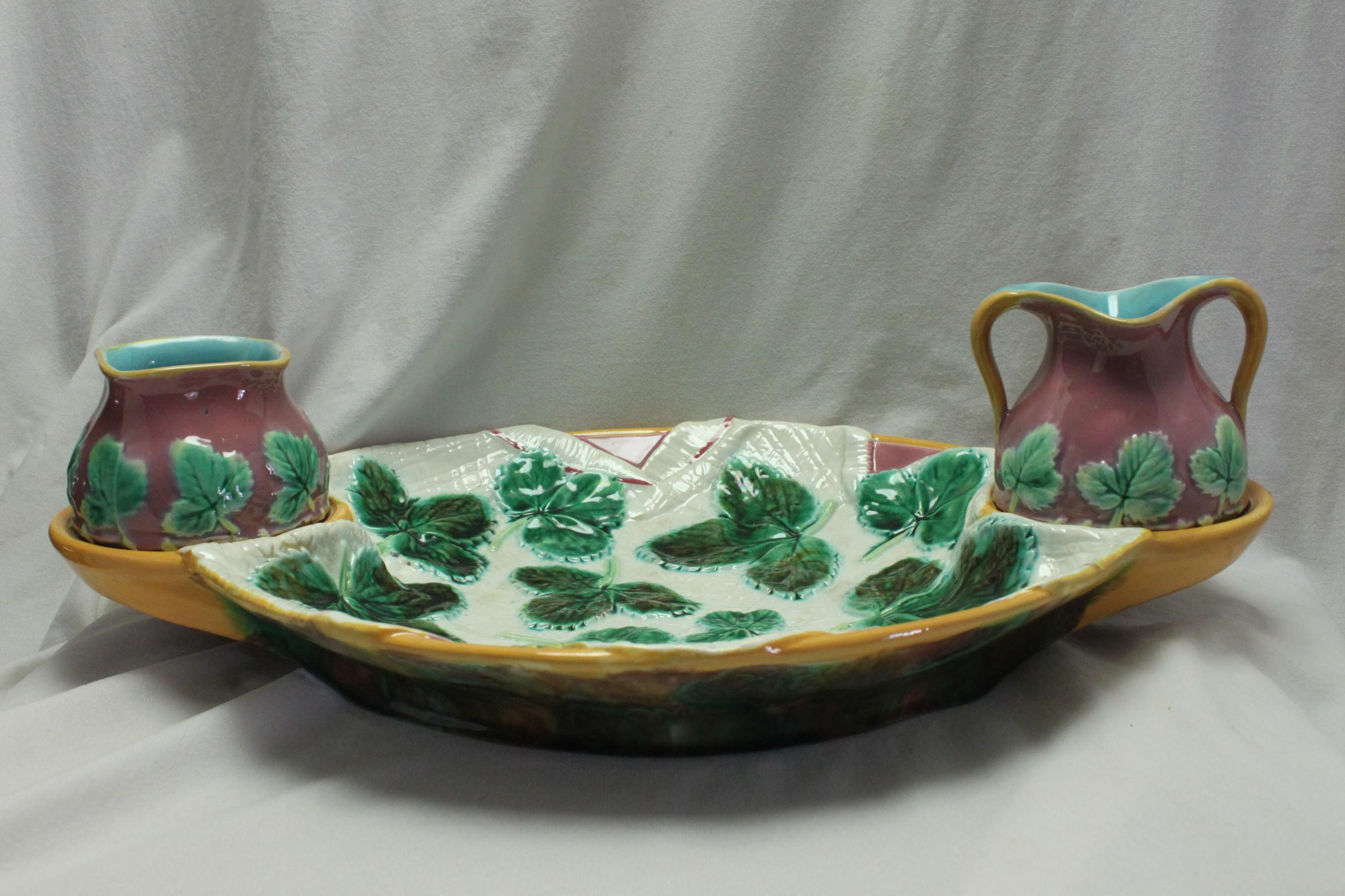 This three piece majolica strawberry set by George Jones features an oval tray with open rings at the ends which hold a small pot of sugar and one of cream. The centre is decorated with a trompe l'oeil effect of a folded napkin scattered with