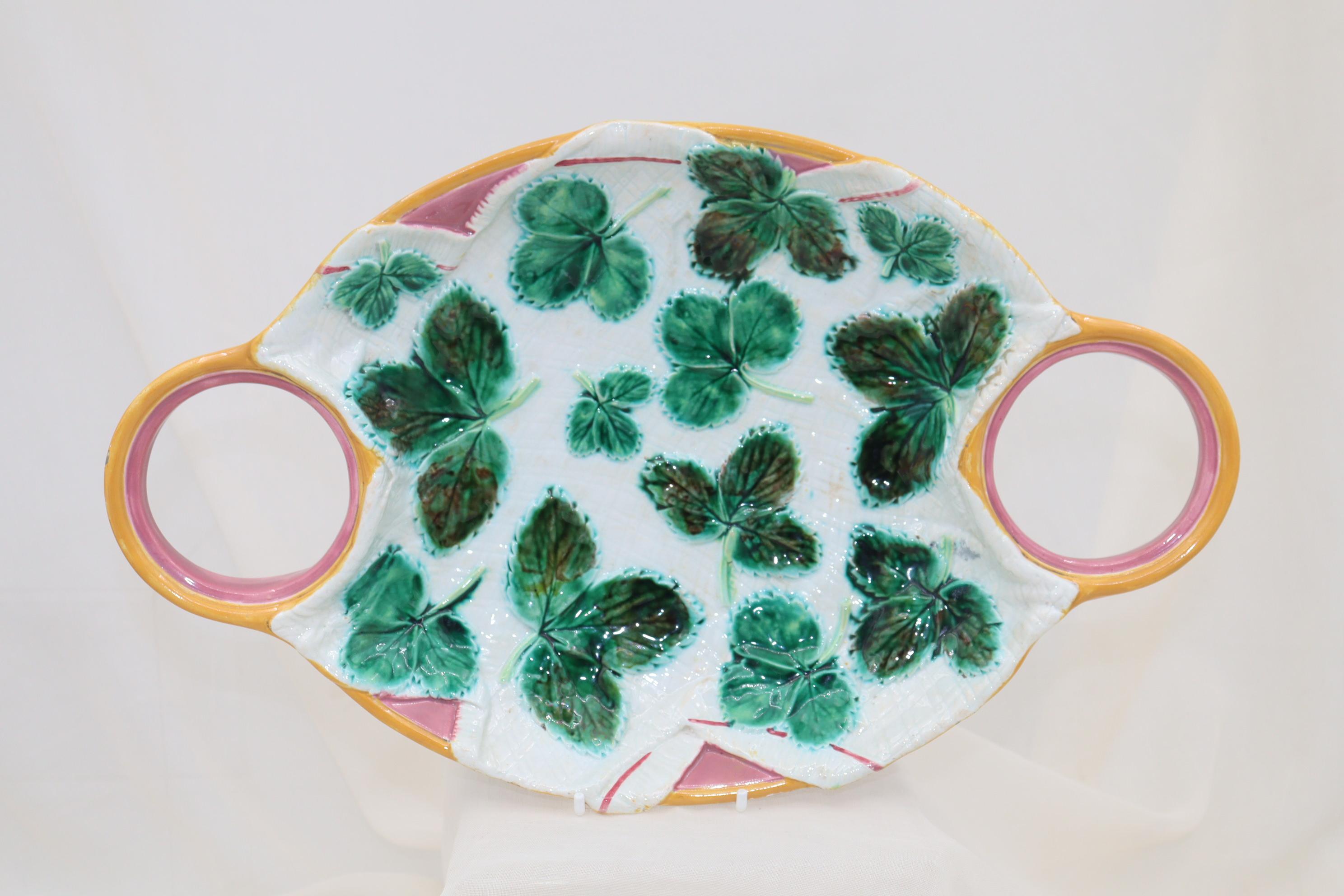 This three piece majolica strawberry set by George Jones & Co. features an oval tray with open rings at the ends which hold a small pot for sugar and one for cream. The centre is decorated with a trompe l'oeil effect of a white folded napkin