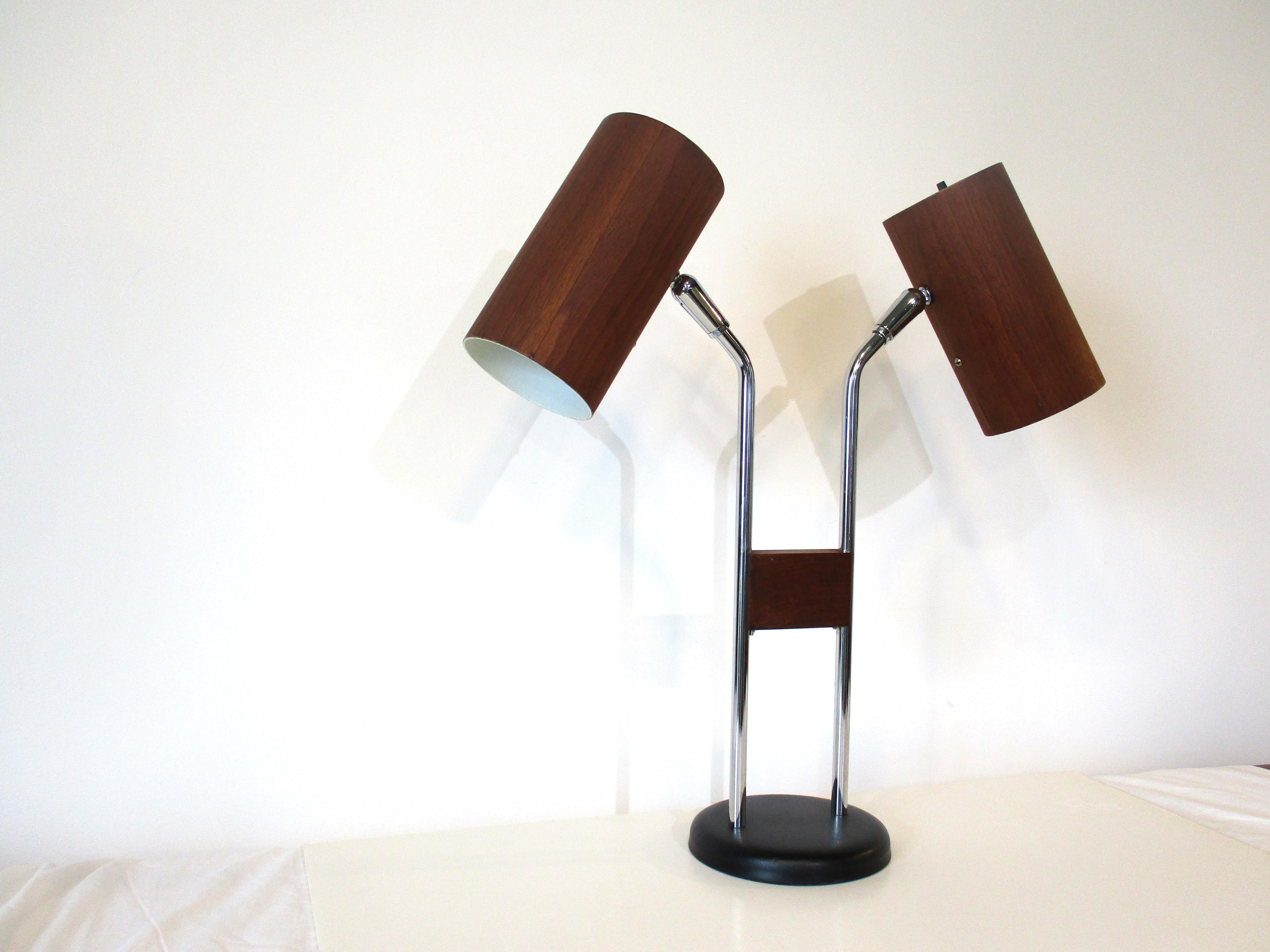 A double headed adjustable table / desk lamp with walnut veneer on both shades . A solid square walnut piece attaches to the center between the chromed poles which are mounted in a satin black steel base . The switches for each light are found on