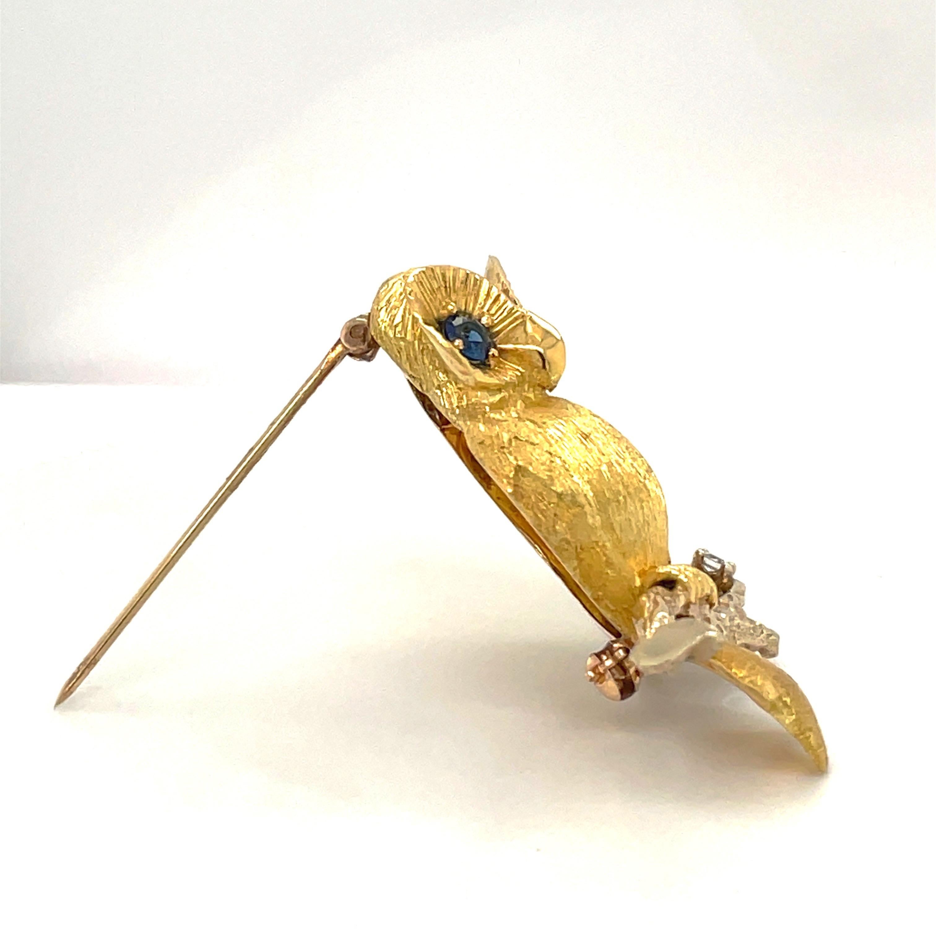 A beautifully sculpted 18 karat yellow gold owl brooch. The sandblasted gold owl sits on a high polished branch tipped with a round diamond. His large eyes are set with round blue sapphires. Stamped 18K.
Made by George Lederman