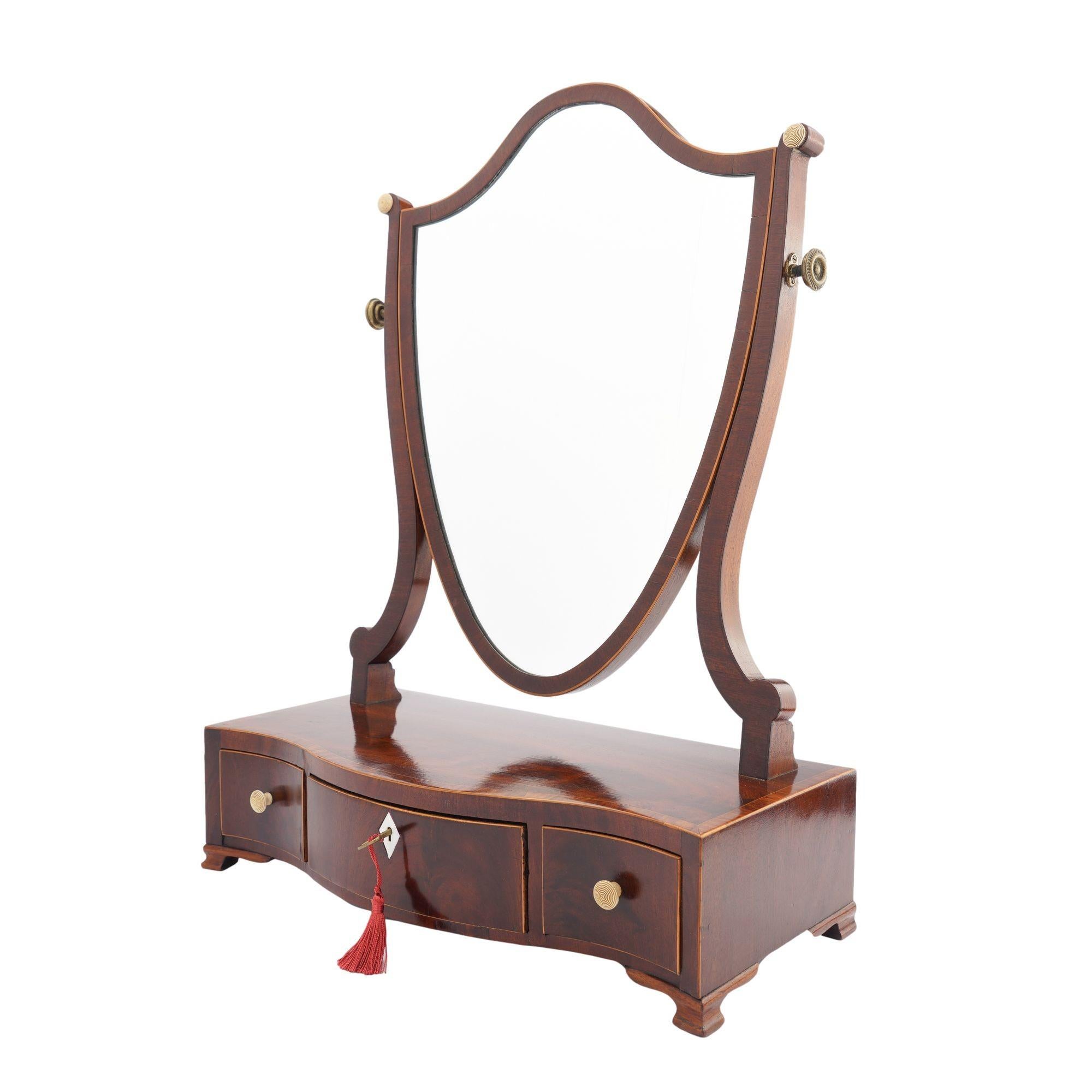 Mahogany swinger shield dressing mirror on a serpentine box stand with divided drawer and ogee bracket feet. The center drawer is fitted with an bone key escutcheon and is flanked by smaller drawers each fitted with turned bone knobs. The shield
