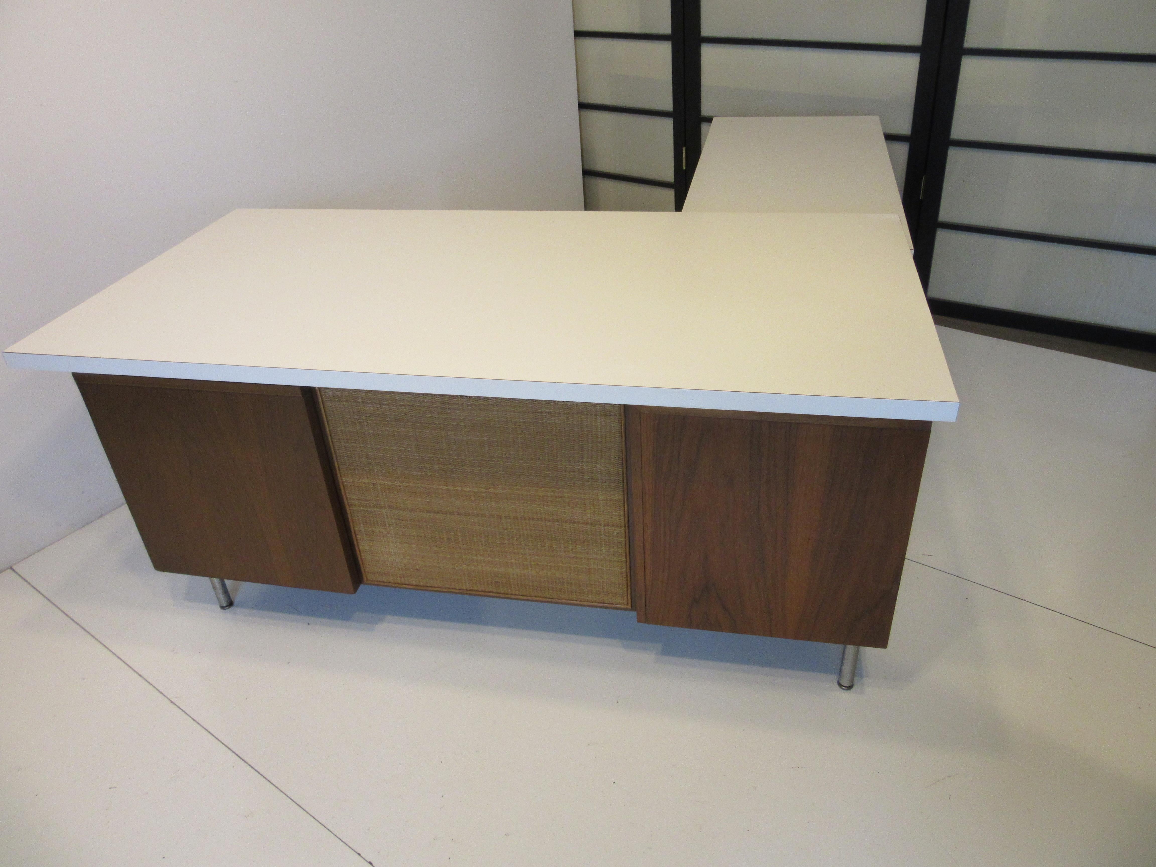 A nice compact sized two piece Nelson desk and return with white laminate floating top, walnut banks with drawers having aluminum J pulls and woven cane privacy screen to the knee hole. The return bolts into the desk and has storage shelves to the