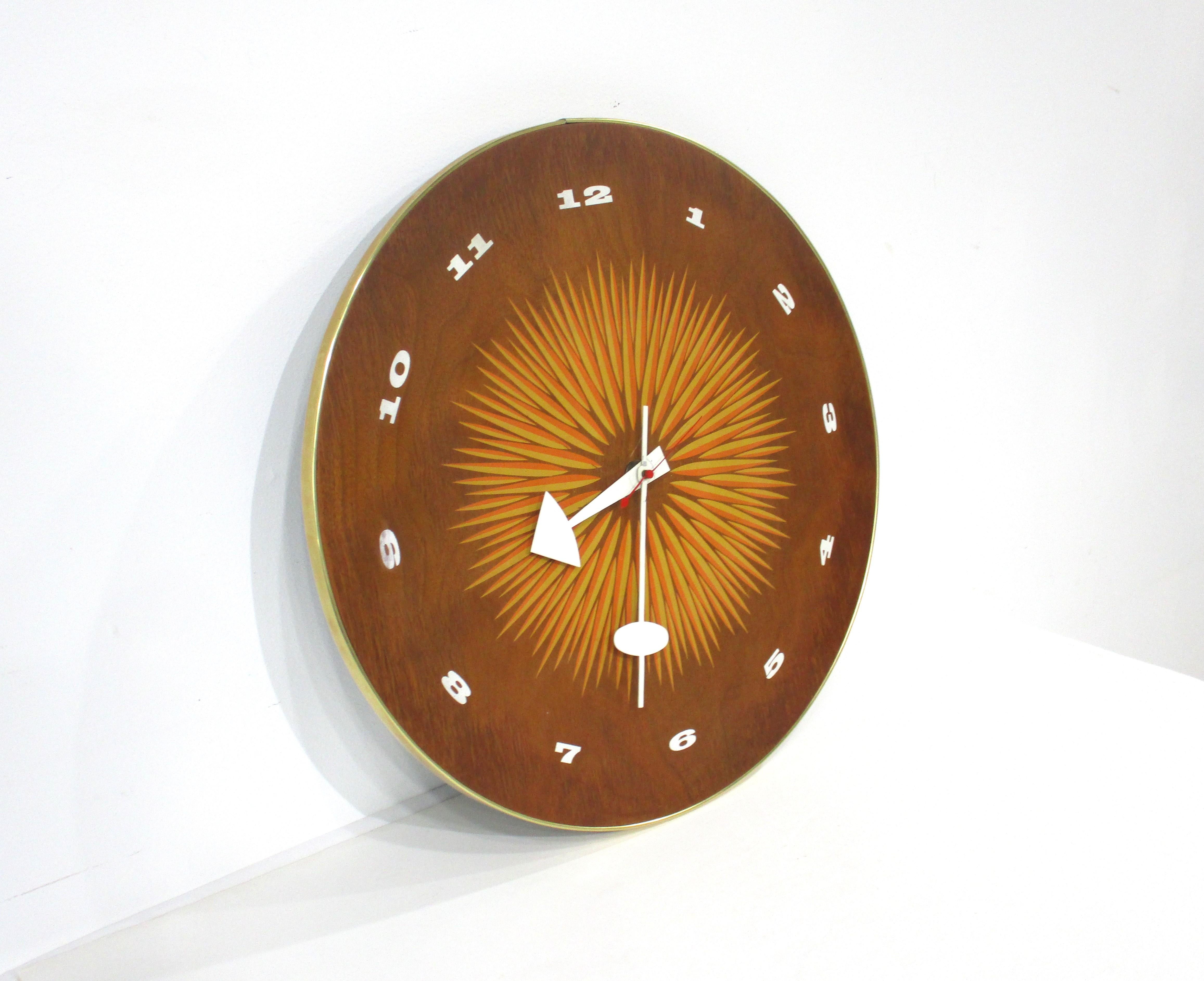A round walnut electric wall clock with printed face having white numbers and a bold sun or flower styled orange and yellow design back round . White metal sculptural hands and red second hand with banded outside edge in a brass toned material .