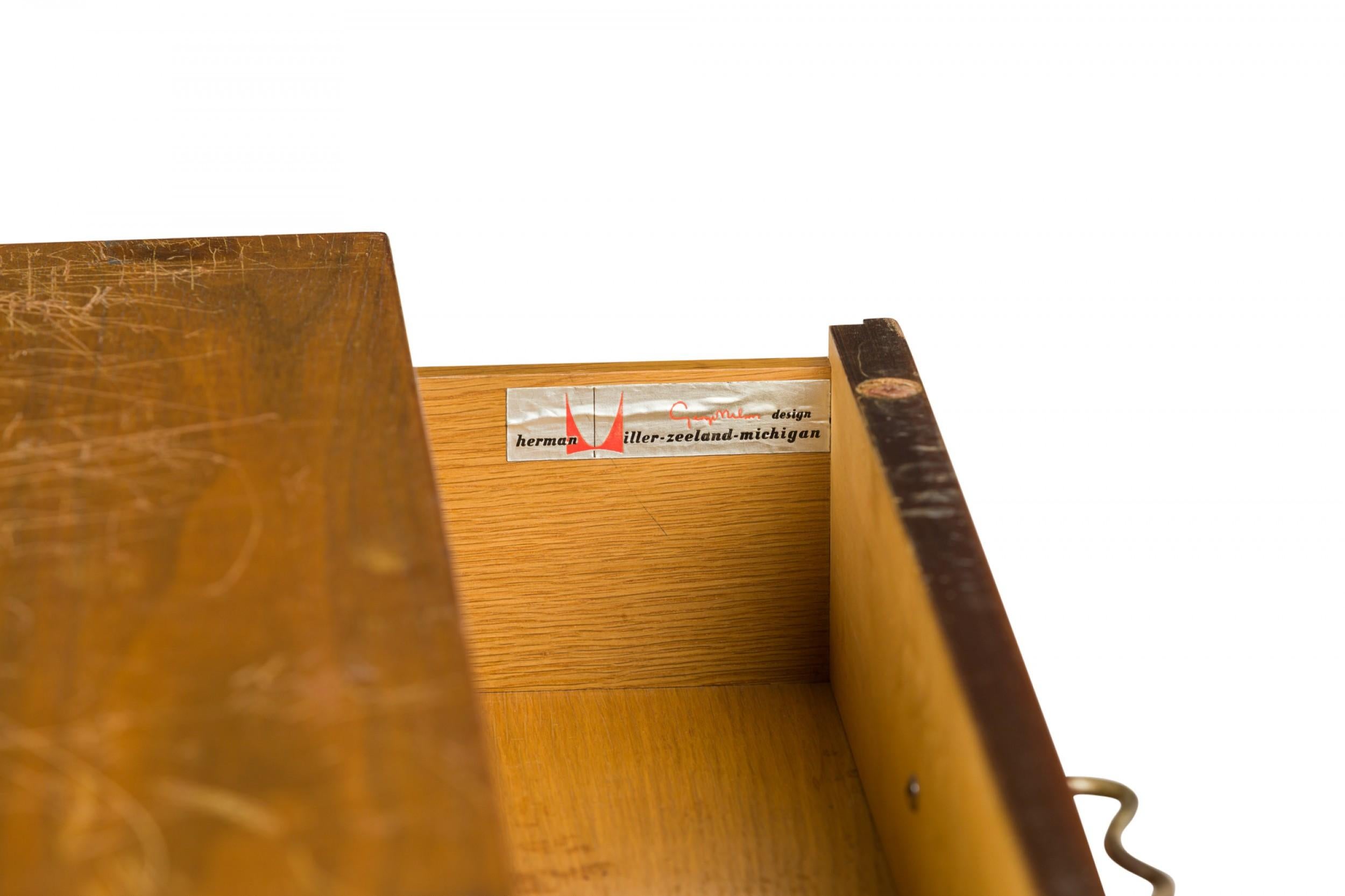 American Mid-Century 4-drawer walnut veneer bedside table / commode with M-shaped silver metal handles, resting on four square wooden legs. (BCS BASIC CABINET SERIES, GEORGE NELSON FOR HERMAN MILLER)(Similar piece: DUF0317B)

Condition: Good; Wear
