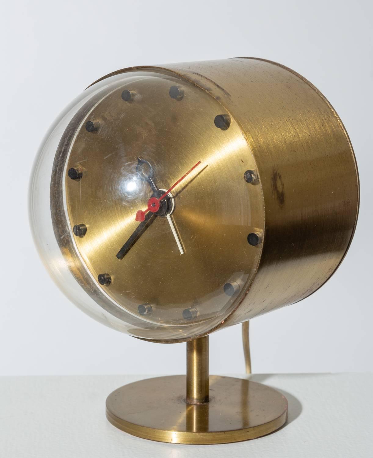 A round brass clock on a brass pedestal foot with a plexiglass bubble case, designed by George Nelson for Howard Miller. The clock has black hour and minute hands, a red second hand, and a white hand for the alarm function. Small black cylinders