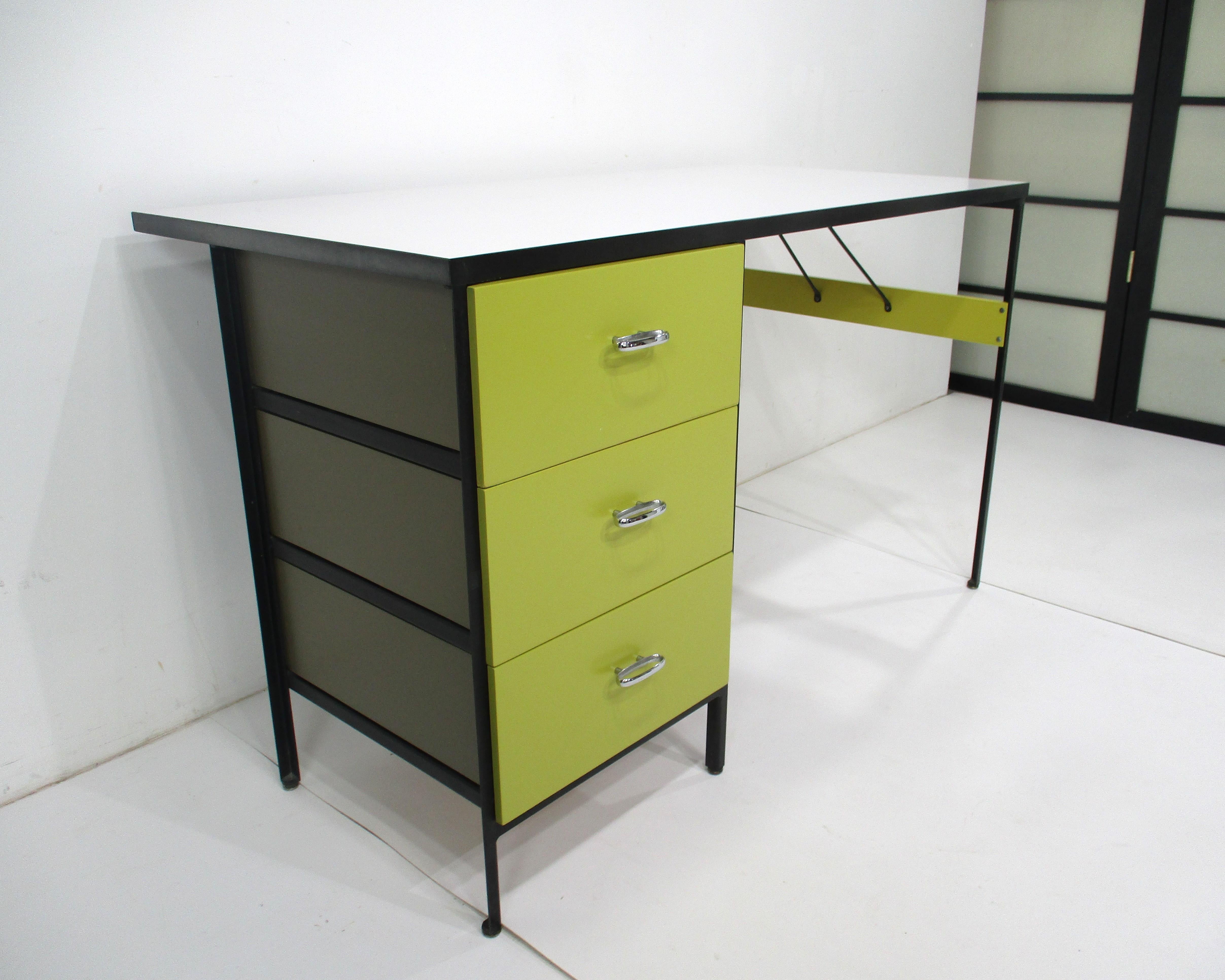 A satin black steel framed desk with okra yellow toned drawer fronts and olive colored sides and backs of drawers . Detailed chrome oval pulls and having a white Laminate desk top giving the piece a perfect Mid Century vibe . Designed by George