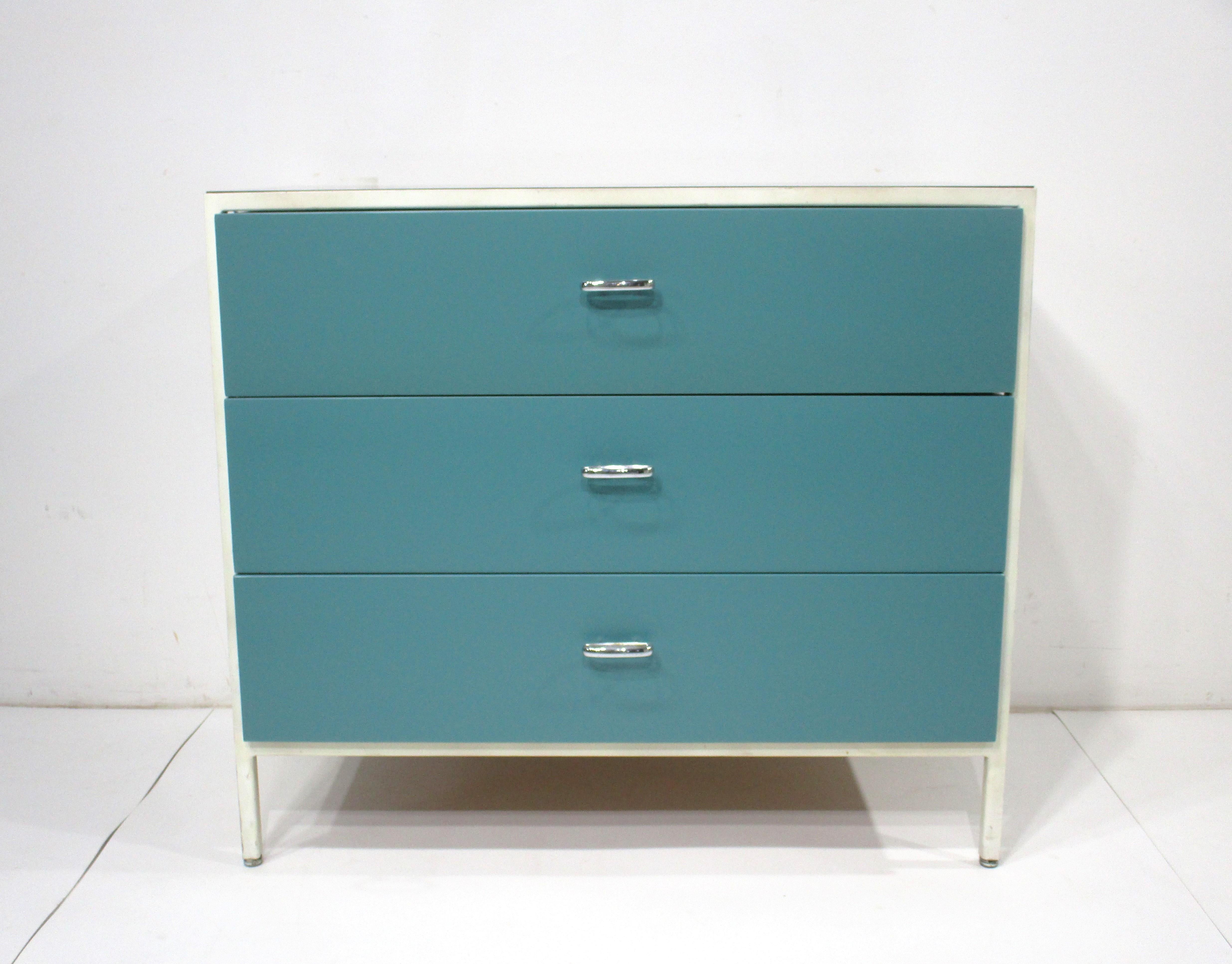 A steel framed dresser chest with off white frame ,  blue fronted wooden drawers having the sides and back painted in contrasting olive green . The oval chrome metal pulls and satin black Laminate top accent the design sensibility of this classic