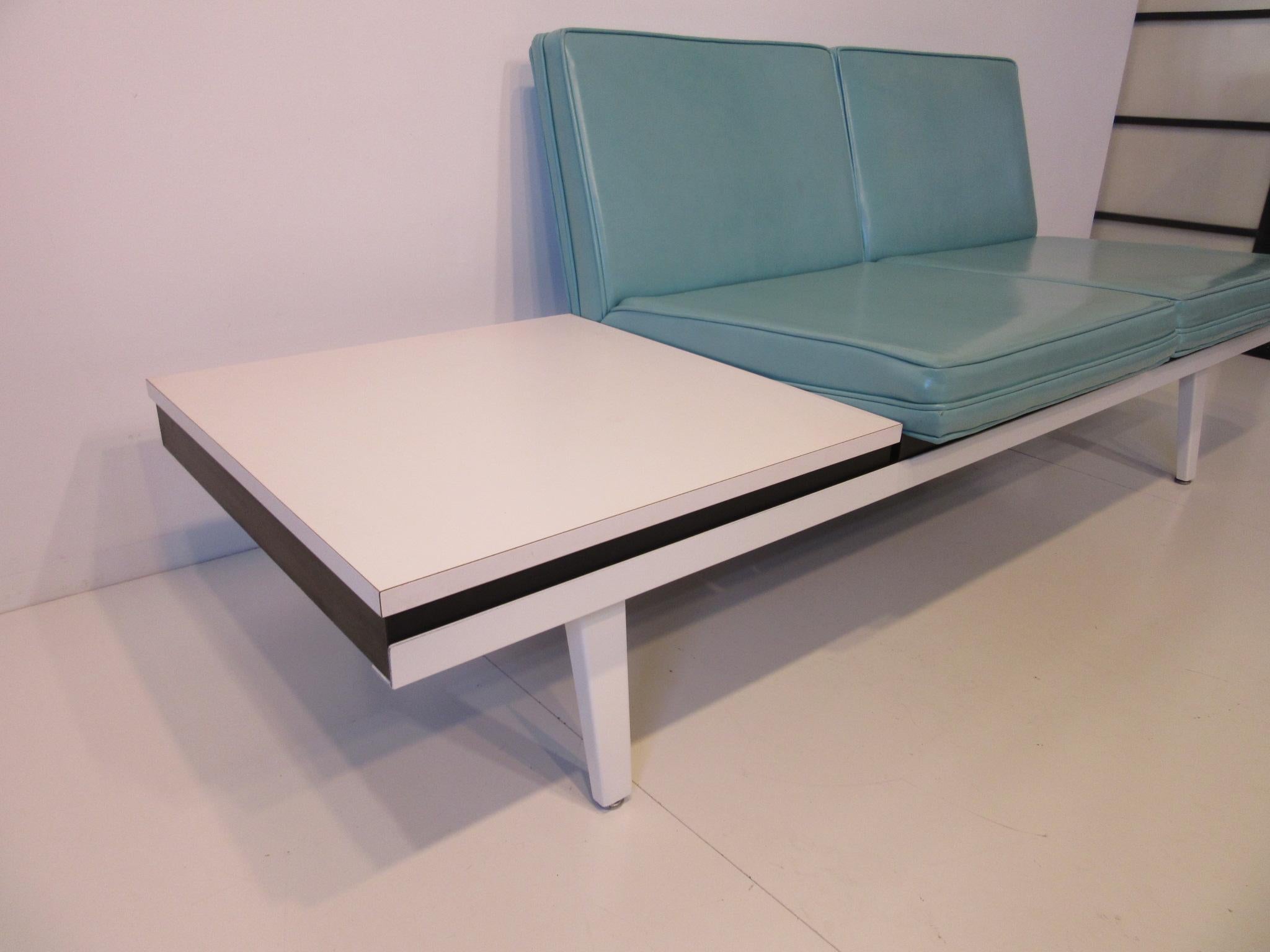 A stain white steel frame Nelson sofa with matching built in side table with white Formica top and wood frame, two turquoise Naugahyde seat cushions sitting on rubber and metal adjustable foot pads. Manufactured by the Herman Miller furniture