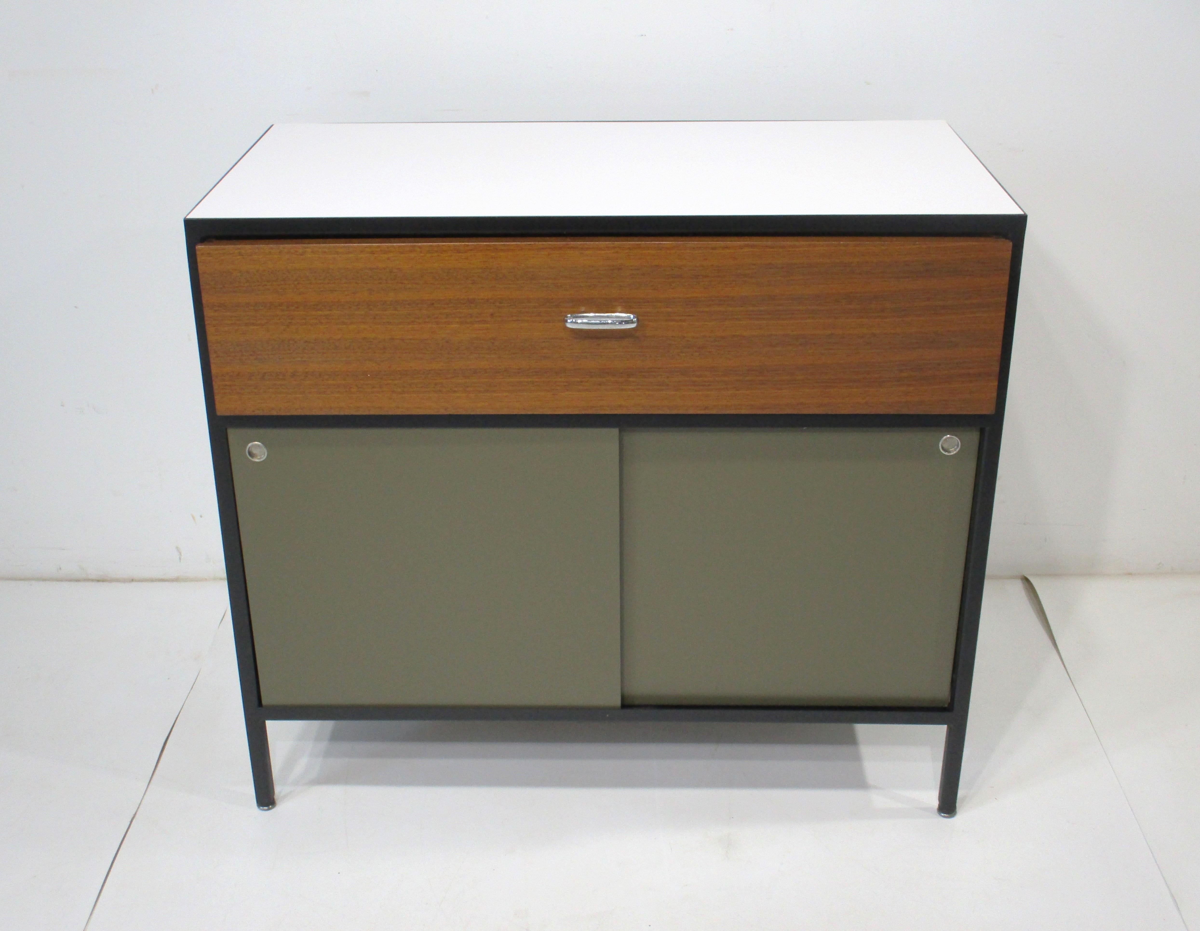 Una cassettiera Steelframe in stile architettonico con struttura in metallo nero satinato, cassettiera e schienale verde oliva e piano in laminato bianco. La parte frontale del cassetto è in noce ben rifinita con un tiretto ovale in metallo cromato,