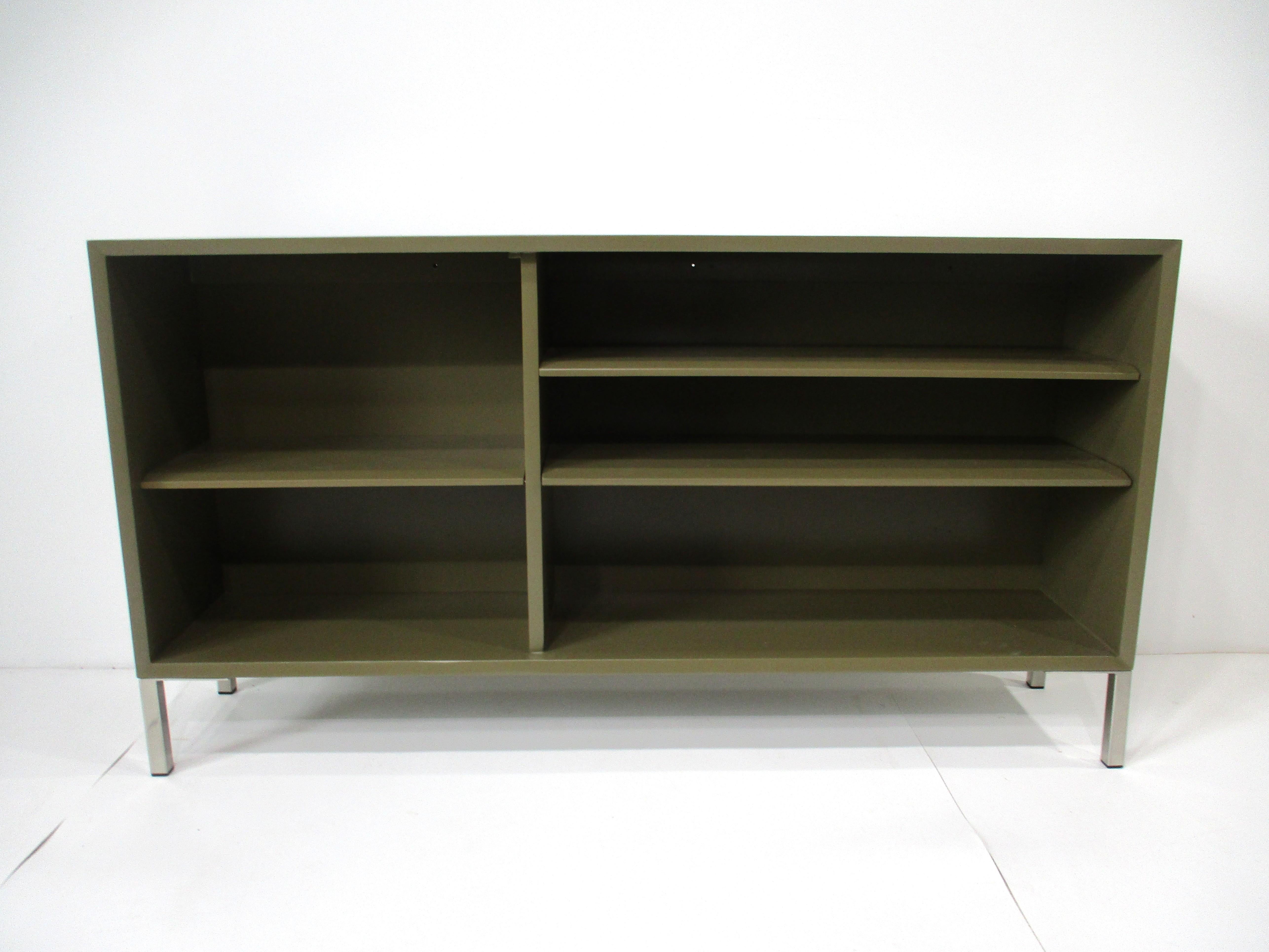 A sturdy wooden bookcase in a olive green finish with three adjustable shelves sitting on stainless steel square tube legs with plastic inserts to protect your floors . This Mid Century piece is designed in the manner of George Nelson with front