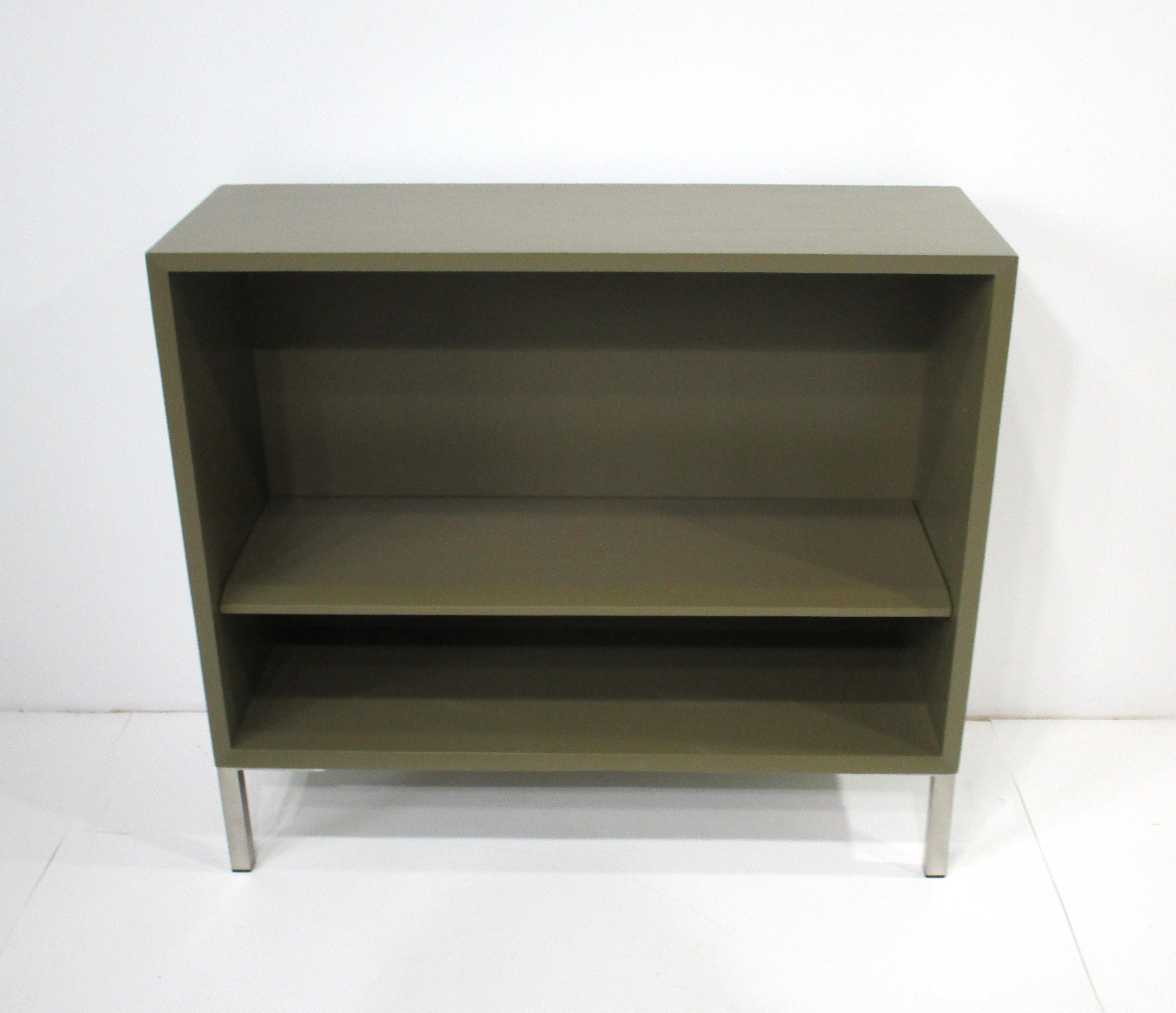 A sturdy wooden bookcase in a olive green finish with one adjustable shelve sitting on stainless steel square tube legs with plastic inserts to protect your floors . This Mid Century piece is designed in the manner of George Nelson with a front