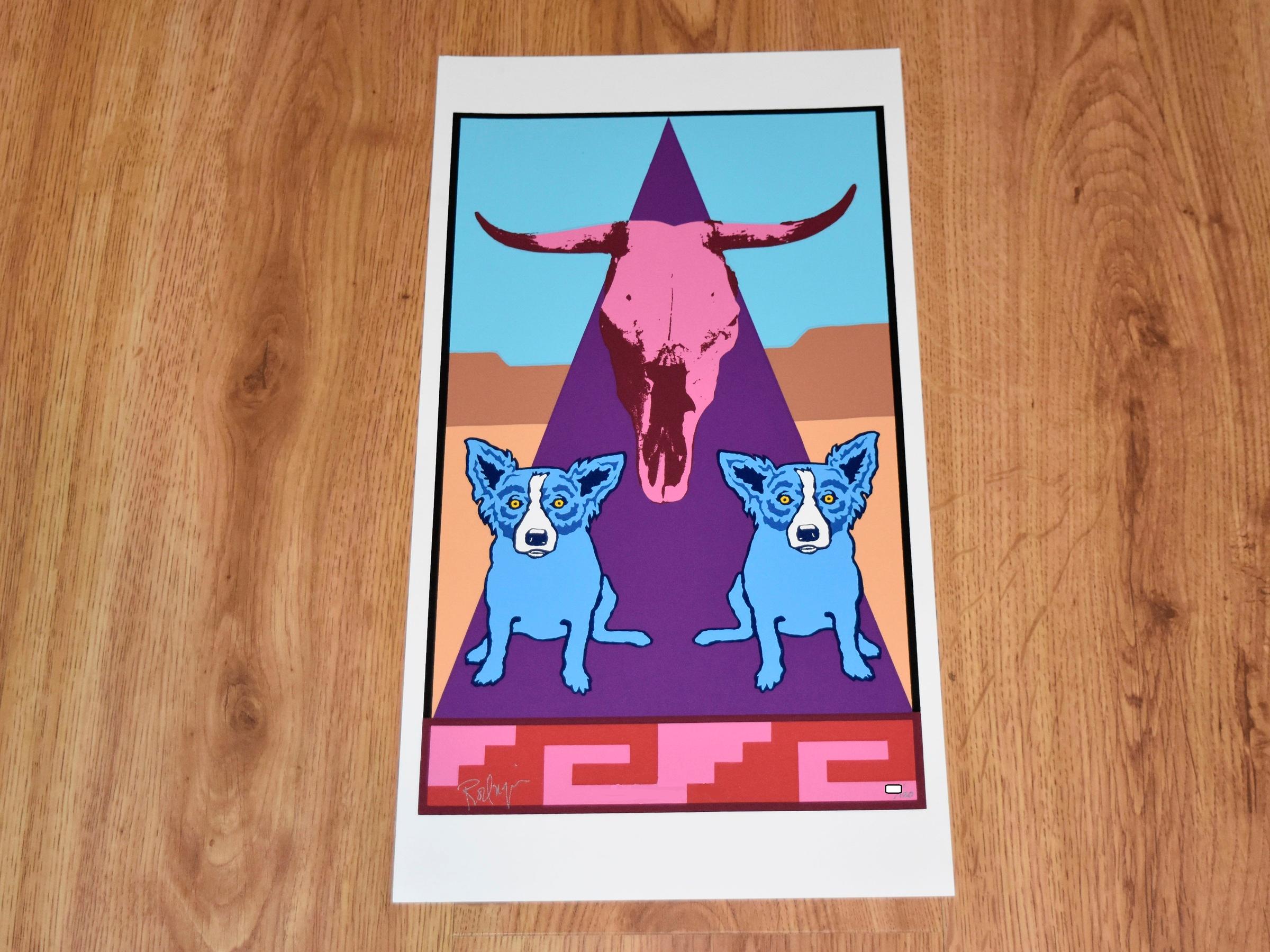 This Blue Dog work consists of 2 blue dogs sitting on either side of a purple triangle with a large steer skull.  The background is of blue sky, desert sand, and mountains in a Southwestern design. At the base of the print is a Southwestern design