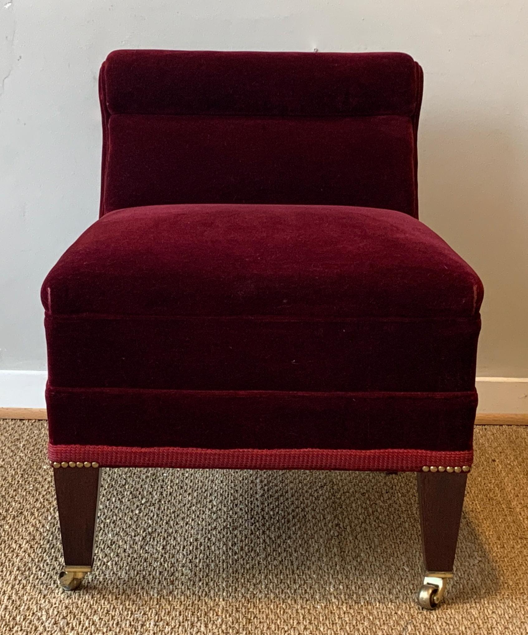 A low slung yet substantial English slipper chair covered in a crimson wool mohair velvet on short tapering legs terminating in brass casters.