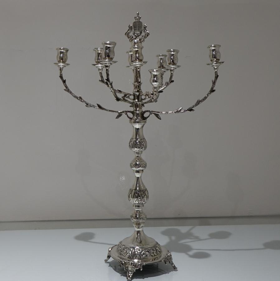 A large silver menorah decorated with sections of ornate embossing to the candlestick. The branch arms are mounted to a spindle and have a elegant naturalistic design. The Shamash and arms are detachable.

 

Measures: Height 22.5