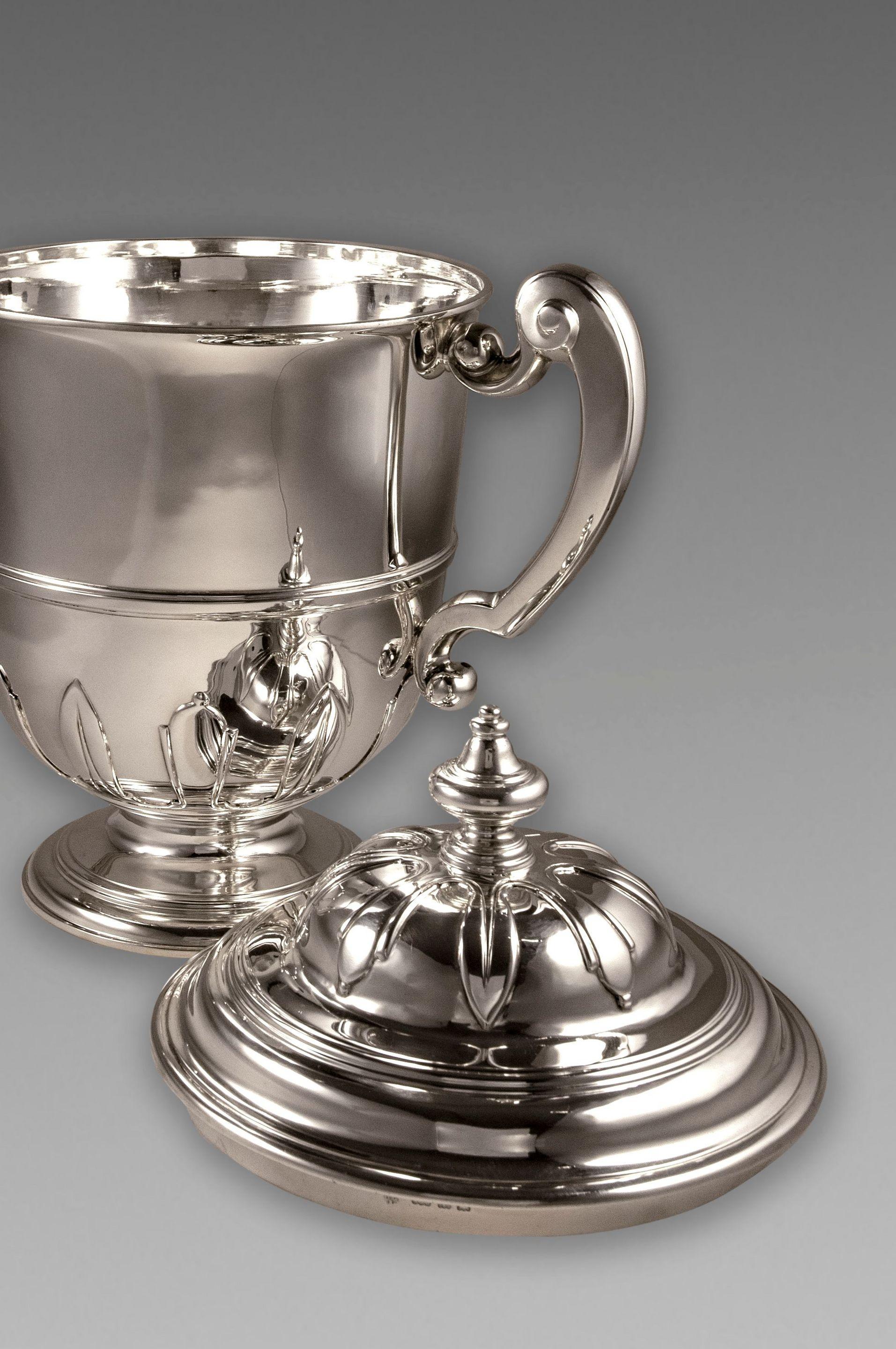 A fine George V sterling silver cup and cover is of a bell-shaped form on to a spreading foot. The surface of the cup is plain and encircled with an applied girdle. The lower body is enriched with an alternating leaf design which is repeated on the