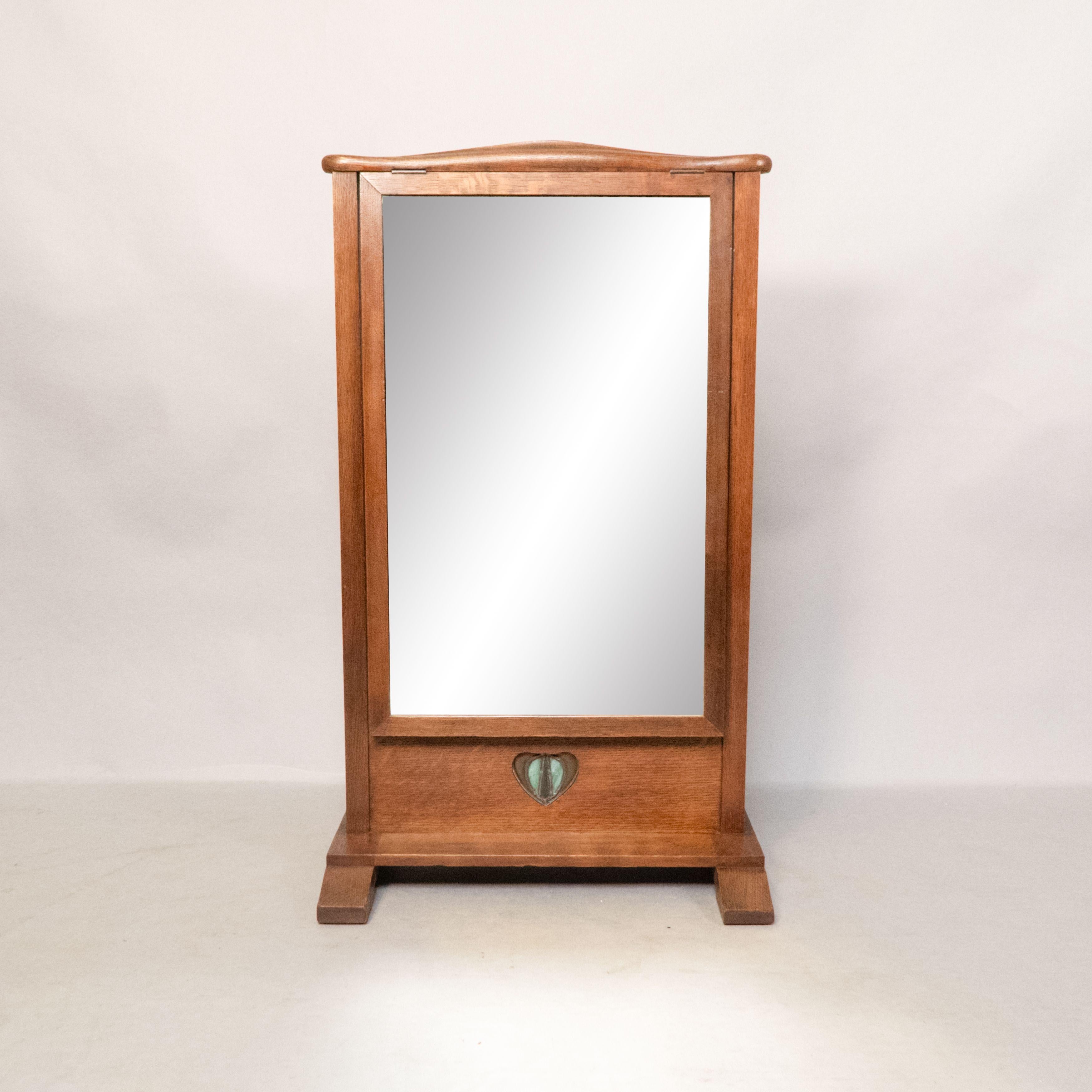 An Arts and Crafts oak dressing table mirror with subtle curved detail to the top and an adjustable mirror on a ratchet mechanism set within it's frame, with a pierced heart to the base centred with a stain glass heart set into copper to form the
