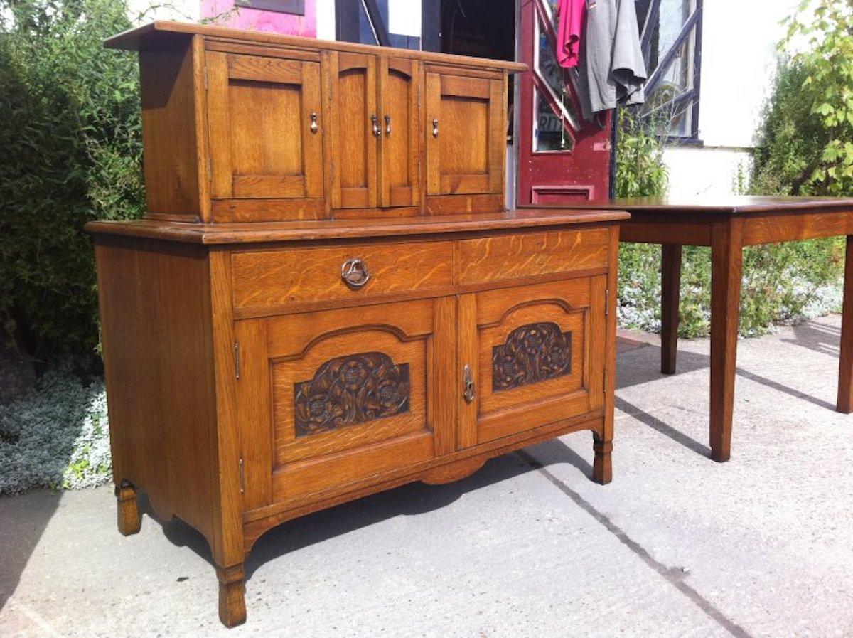 A small Arts & Crafts oak sideboard in the style of George Walton.
With flaring cornice, four small upper cupboards the central pair with arched doors and a small work area in front, a pair of drawers with stylized copper handles (the other handle