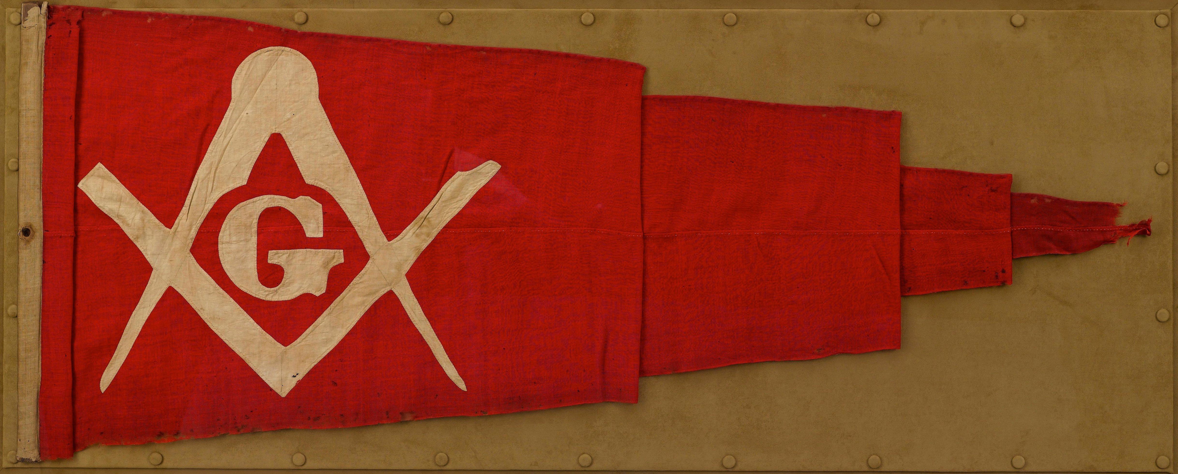 This is a Civil War-era triangular banner with hand-sewn Freemason insignia. The banner is constructed of cotton. It has a rich red field, a hand-sewn white insignia on the hoist end, and a white cotton headband. The banner is from an unknown maker,