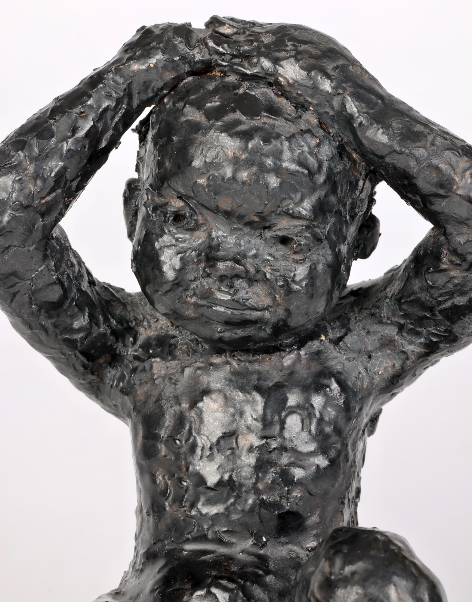 Eine bezaubernde, schwarz glasierte Studio-Keramik-Skulptur eines sitzenden Kindes von George West, datiert 1969. Die Figur ist von Hand aus rotem Ton modelliert und stellt ein sitzendes Kind dar, das seine Hände auf dem Kopf hält. Die Figur hat
