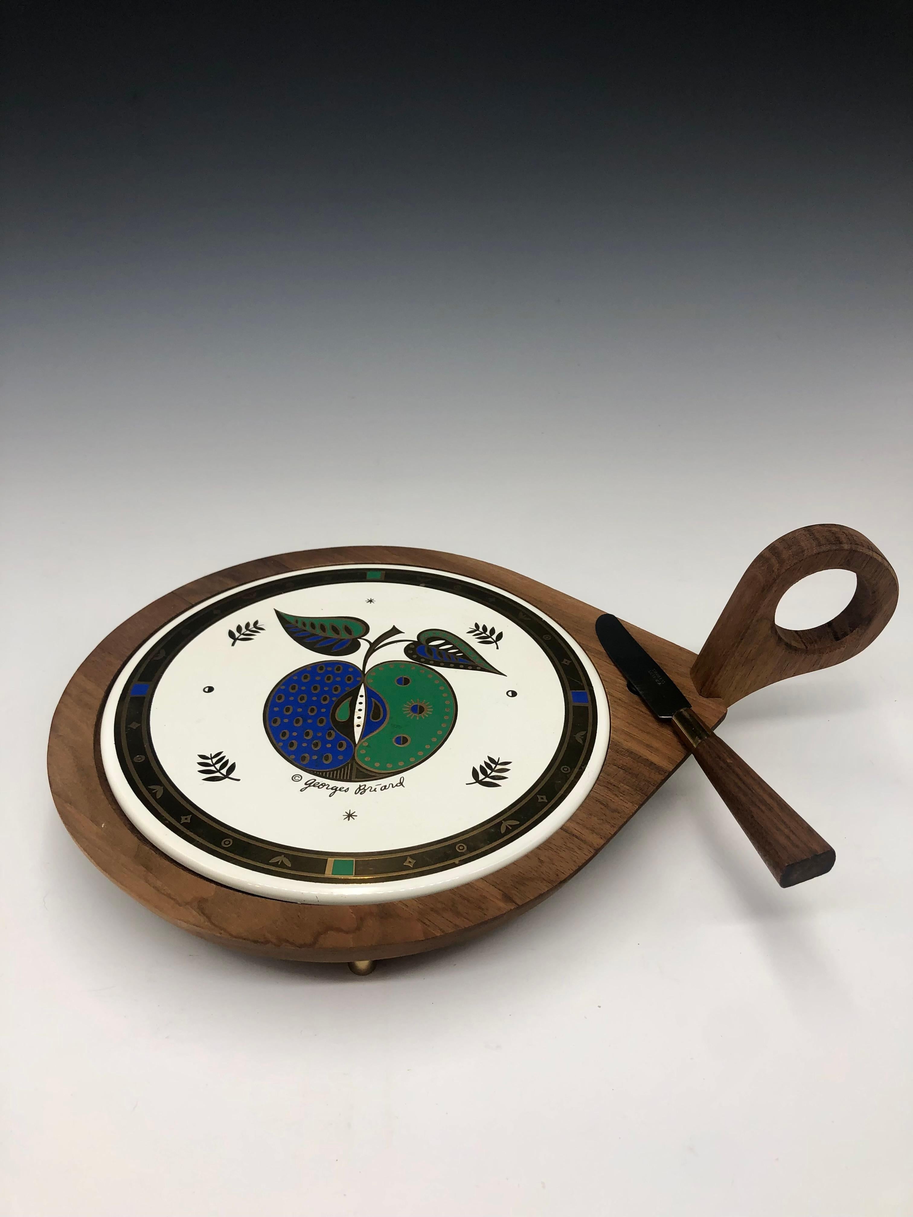 A striking mid-century modern cheese serving board by celebrated designer Georges Briard, featuring his signature bold, graphic style. 

This piece combines a rich, solid walnut frame with a removable round enamel insert decorated in vibrant blues,