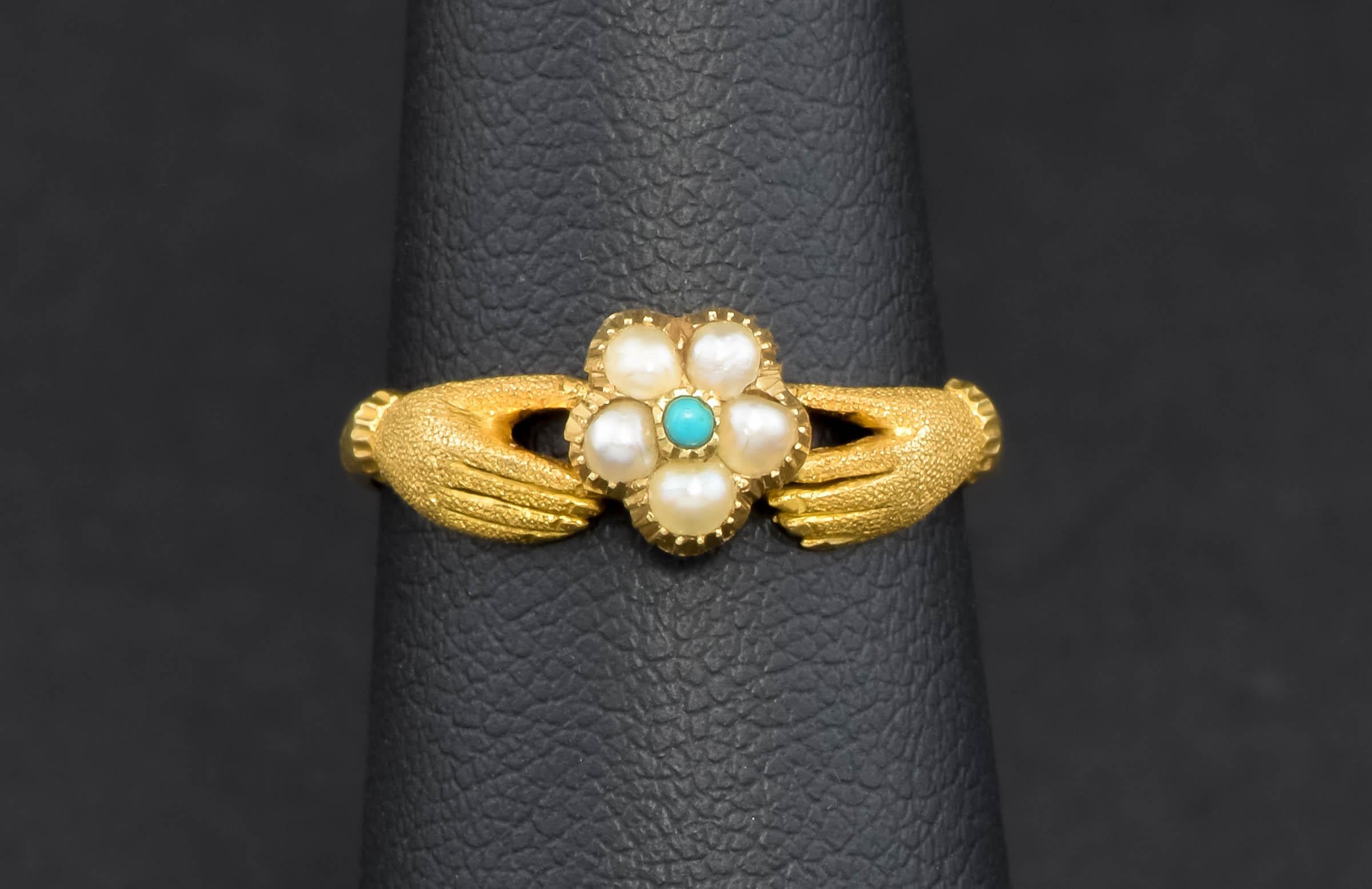 Crafted of 14K gold with a lovely bloomed finish, the ring features a classic design of two cuffed hands holding a flower (a pansy or forget-me-not, most likely, in friendship, love and remembrance.) The flower blossom’s petals are set with pearl,