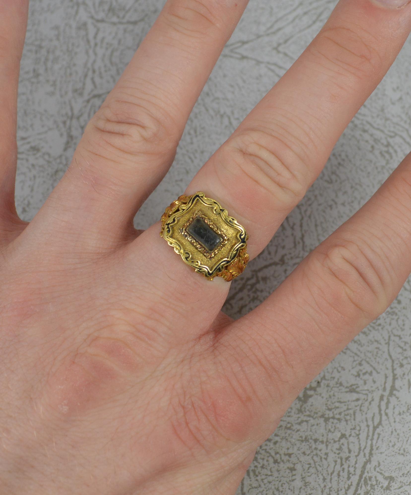 A Georgian era mourning ring.
18 carat yellow gold example. Set with a rectanglar glass panel to centre.
Surrounding is a scroll work border with split floral engraved shoulders.
English hallmarked example.
15mm x 12mm cluster head.

CONDITION ;