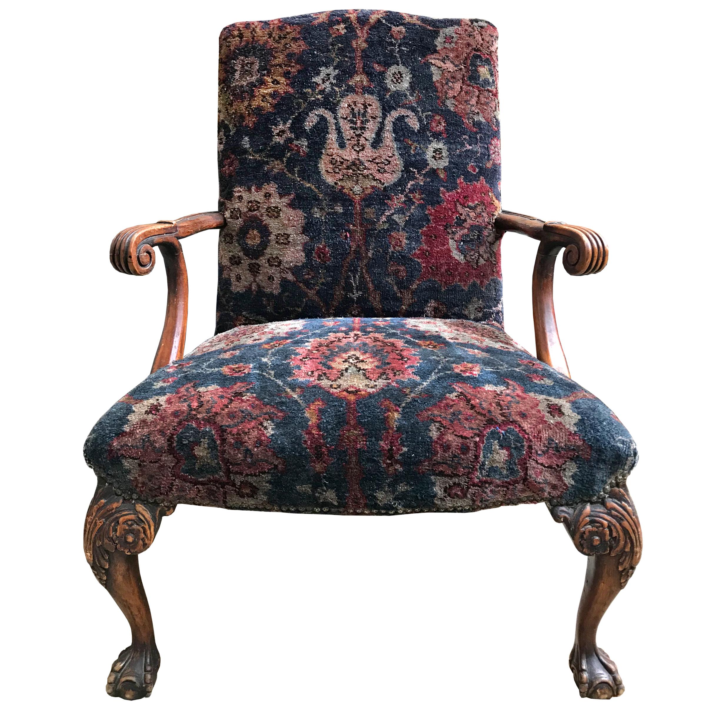 A 19th century Georgian period armchair with curved and scroll carved arms, claw and ball feet, and a wide straight back. The footrest has bun feet. Both have been upholstered in the same 19th century Persian rug, with nailhead trim. 

Dimensions