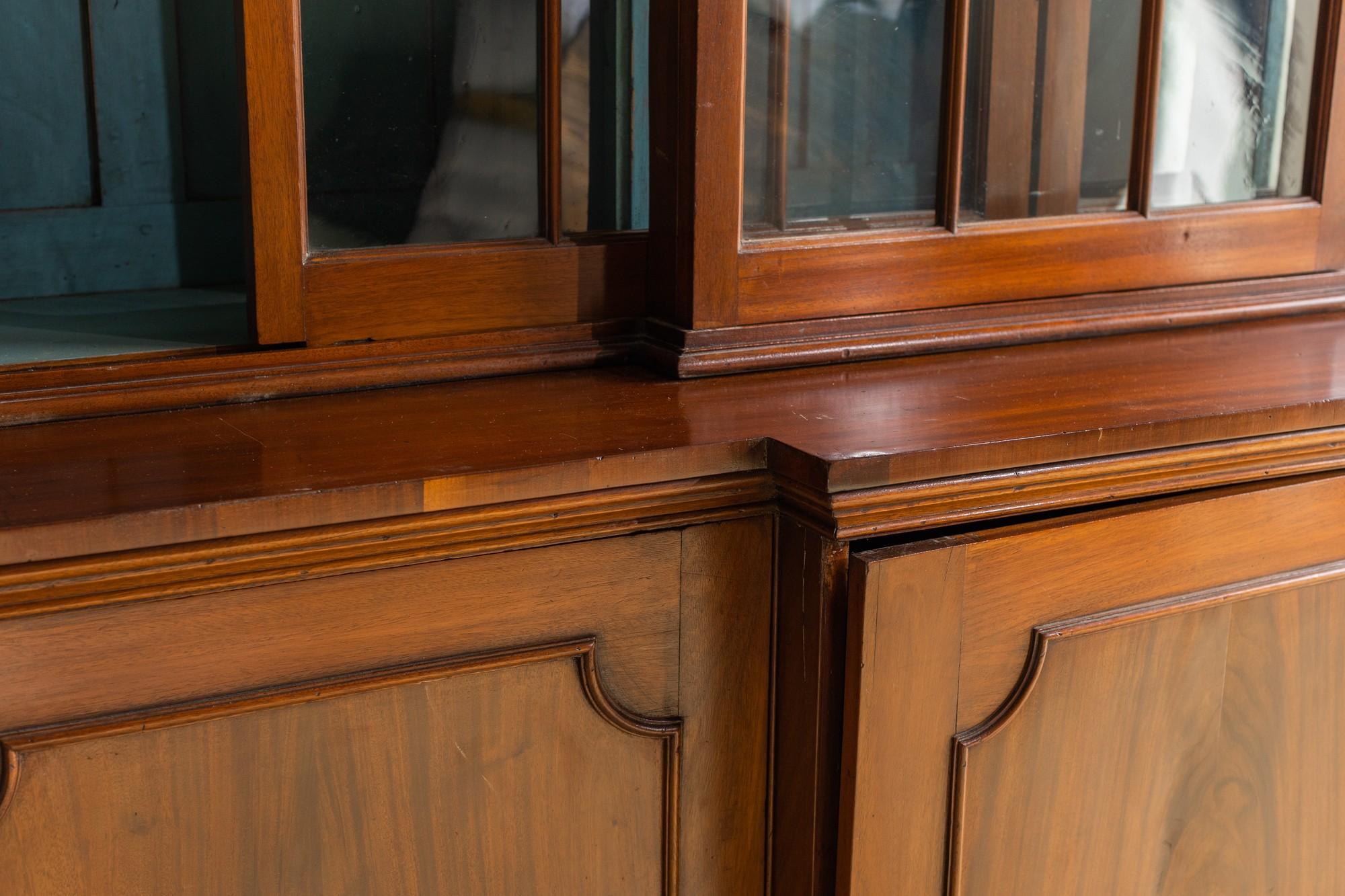 This early 19th century Georgian breakfront bookcase has a beautiful medium brown stain finish on its mahogany wood. The glass is original and there is dentil molding at the crown. The interior of this piece is painted blue with three interior